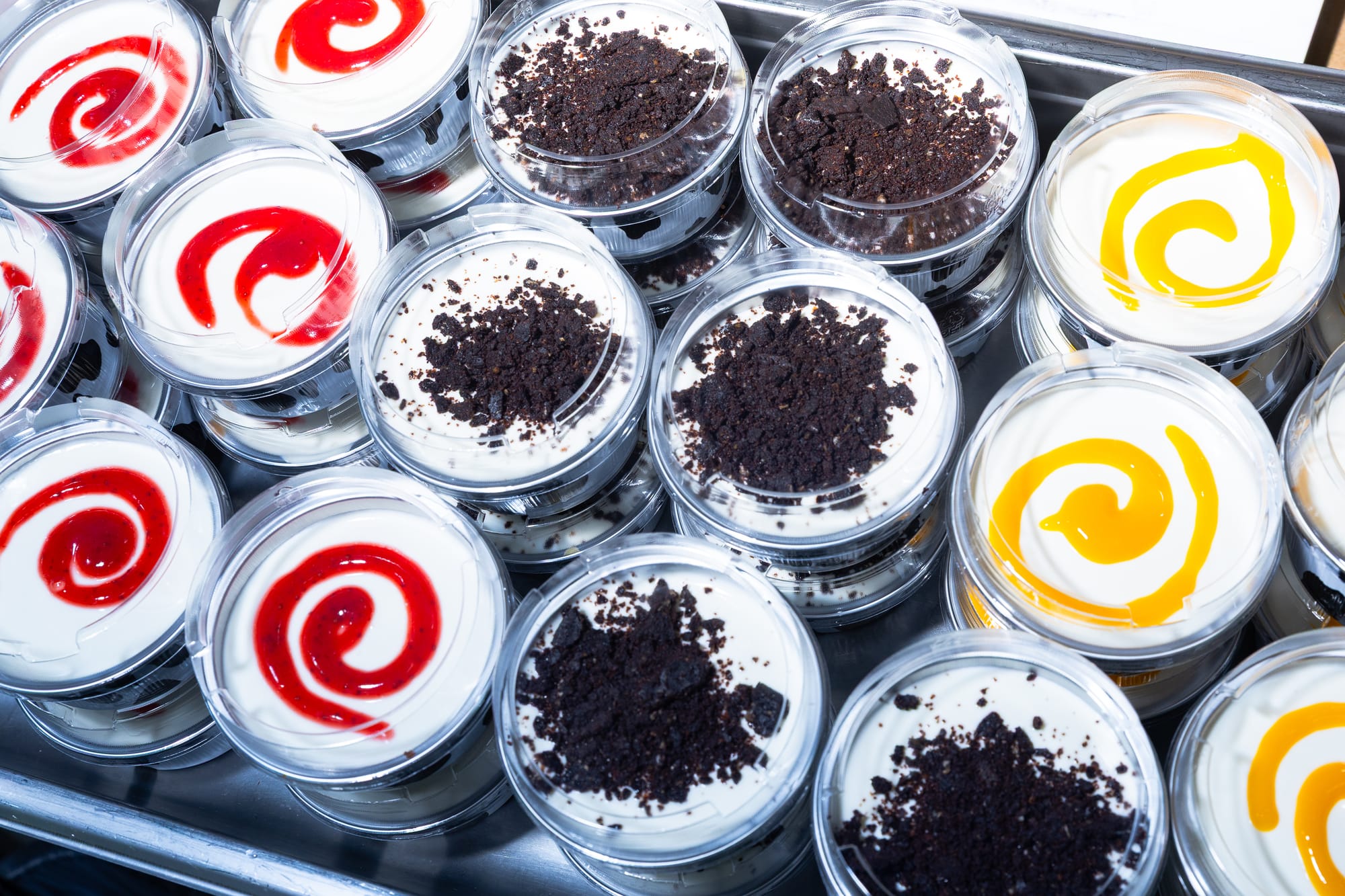 Display of small round cheesecakes. From left to right, with red swirl topping, a crumbly chocolate topping, and a yellow swirly topping.