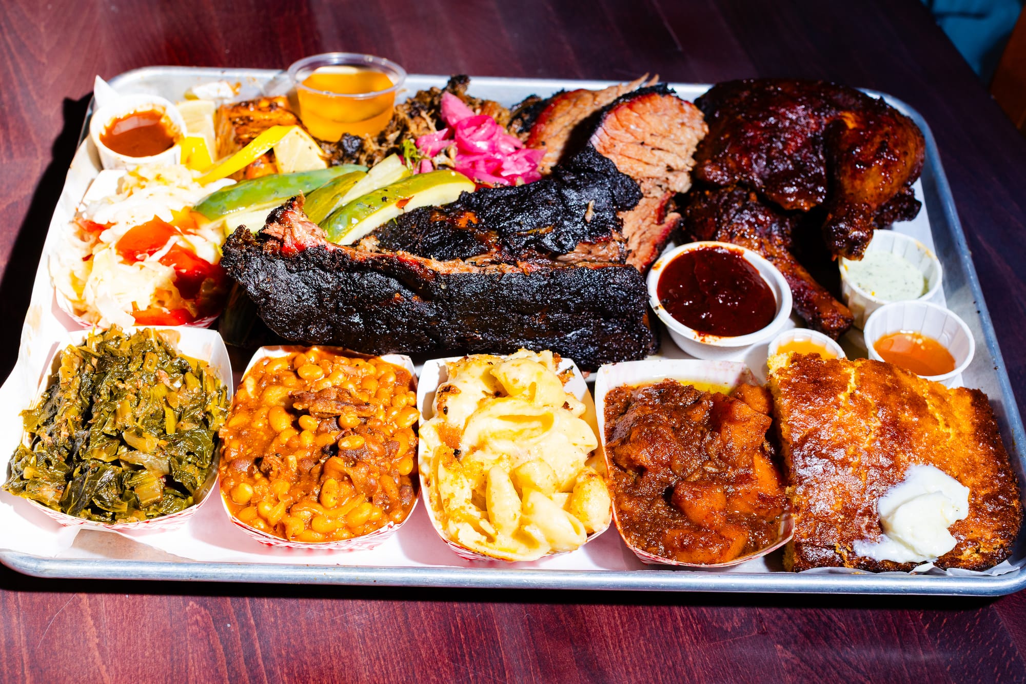Sheet pan of barbecued meats and sides along the bottom. From left to right: collard greens, beans, macaroni and cheese, candied sweet potatoes, and cornbread.
