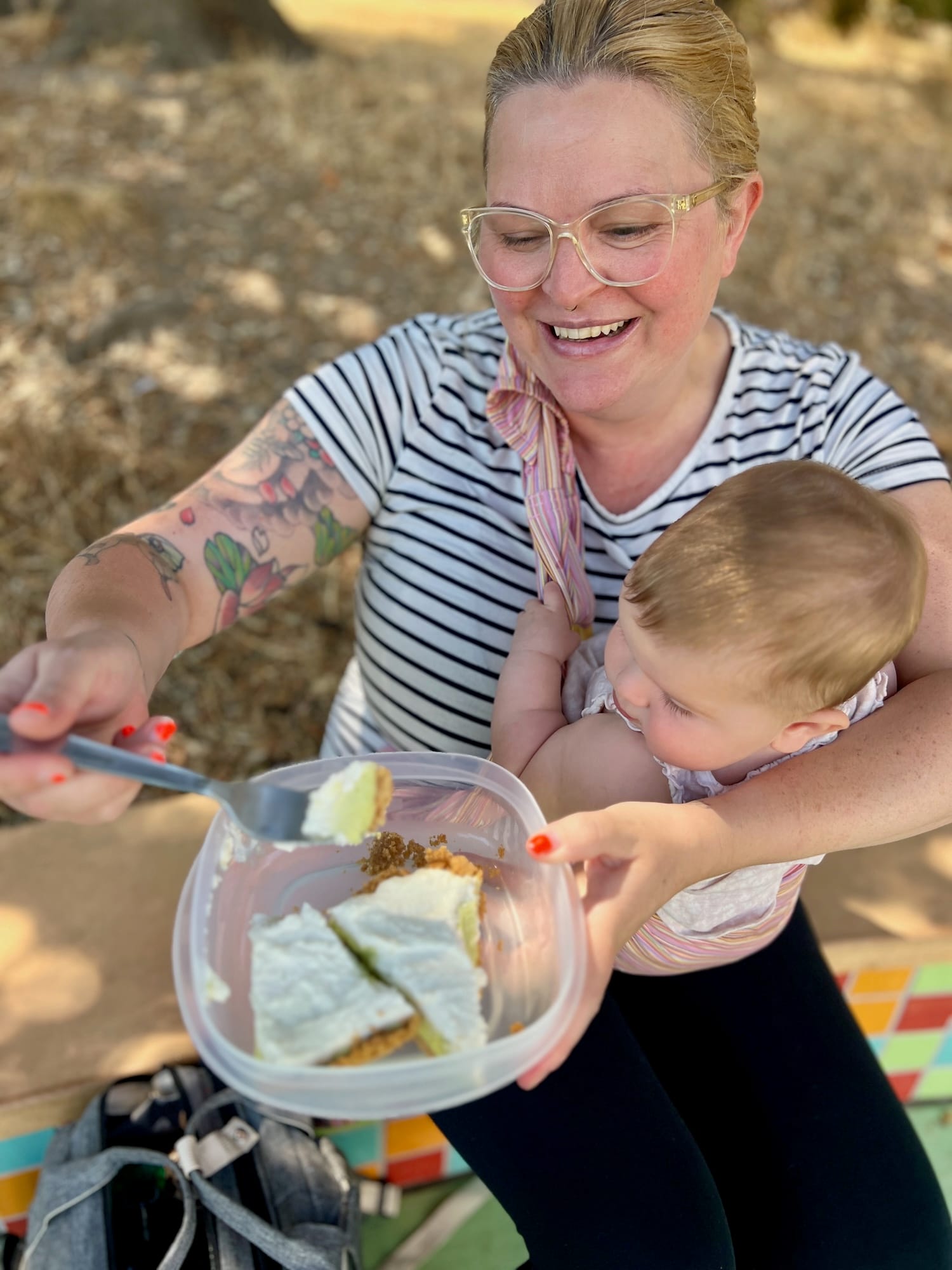 A woman wearing clear glasses smiles and eats a piece of pie out of a tupperware. She has a baby strapped to her chest, who is eyeing the pie with interest.