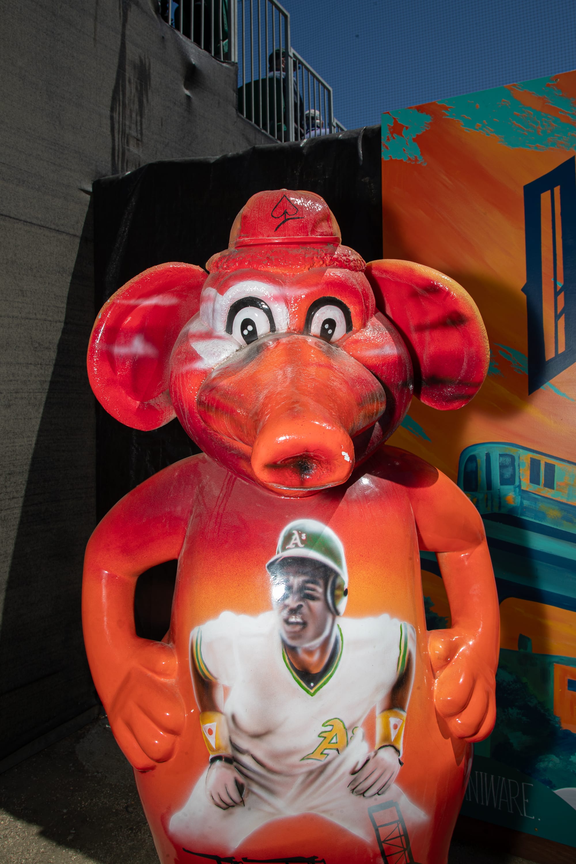Red statue of an anthropomorphic elephant with a painting of a dark-skinned man in a white Oakland A's uniform crouching on its torso.