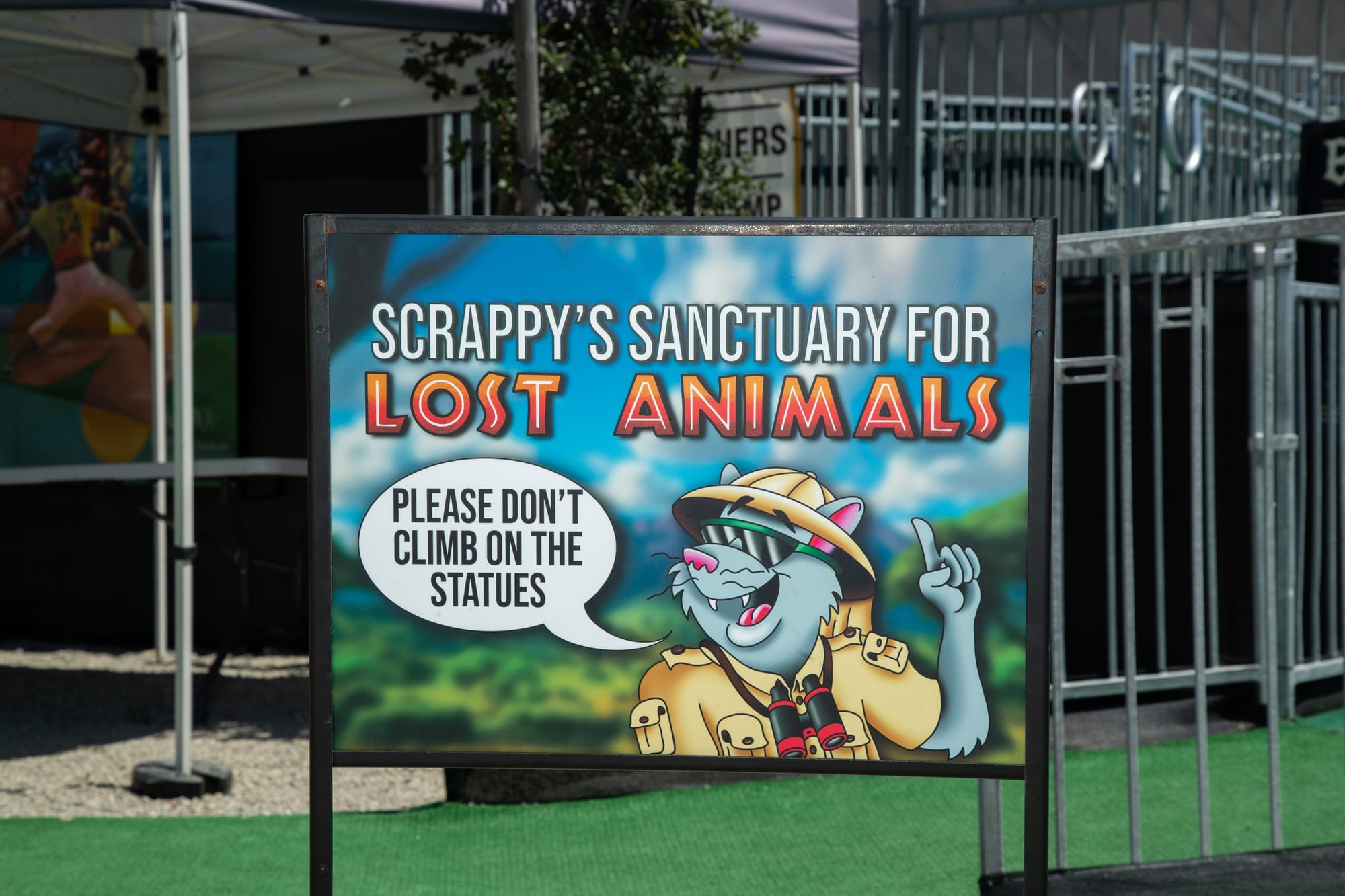 A sign depicting a grey mammal mascot in safari gear. Text says "Scrappy's sanctuary for lost animals." A speech bubble says "Please don't climb on the statues."
