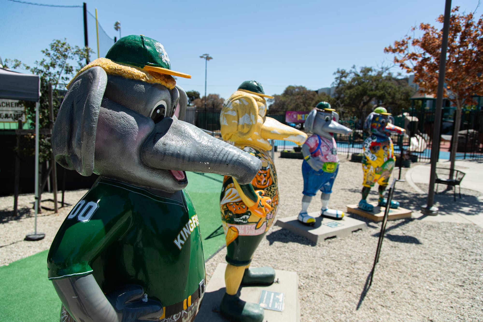 Four statues of anthropomorphic elephants stand in a line in front of a baseball field.