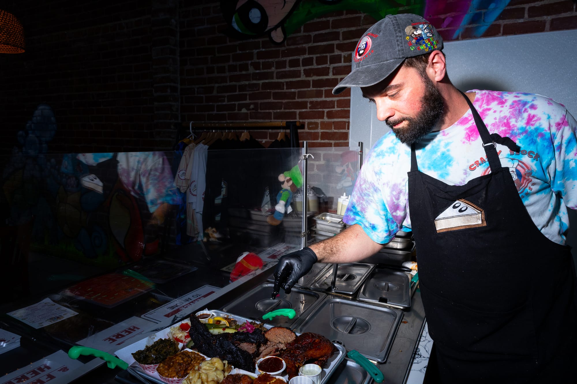 Bearded man in a tie-dyed shirt and black apron gestures toward a sheet pan full of barbecued meats.