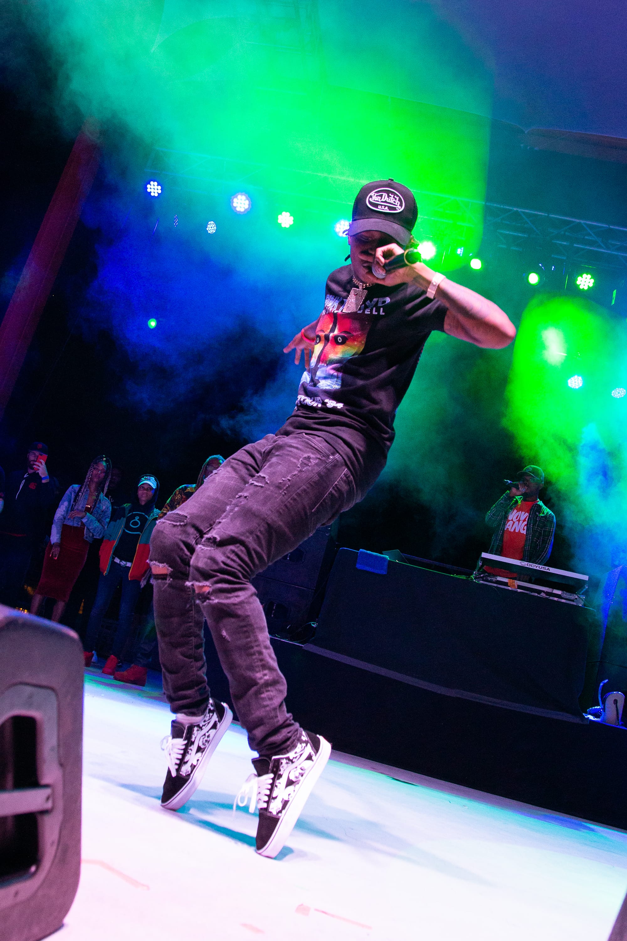 A man in a black t-shirt stands on tiptoes while rapping into a microphone on a stage.