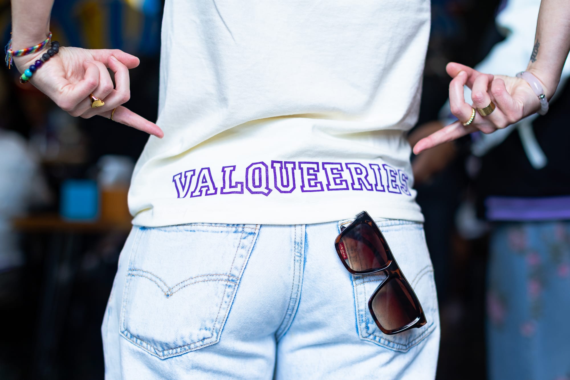 the back of a person wearing light blue jeans and a white shirt with the word 'valkyries' printed in purple and sunglasses in their back pocket; they are pointing at the word with both hands
