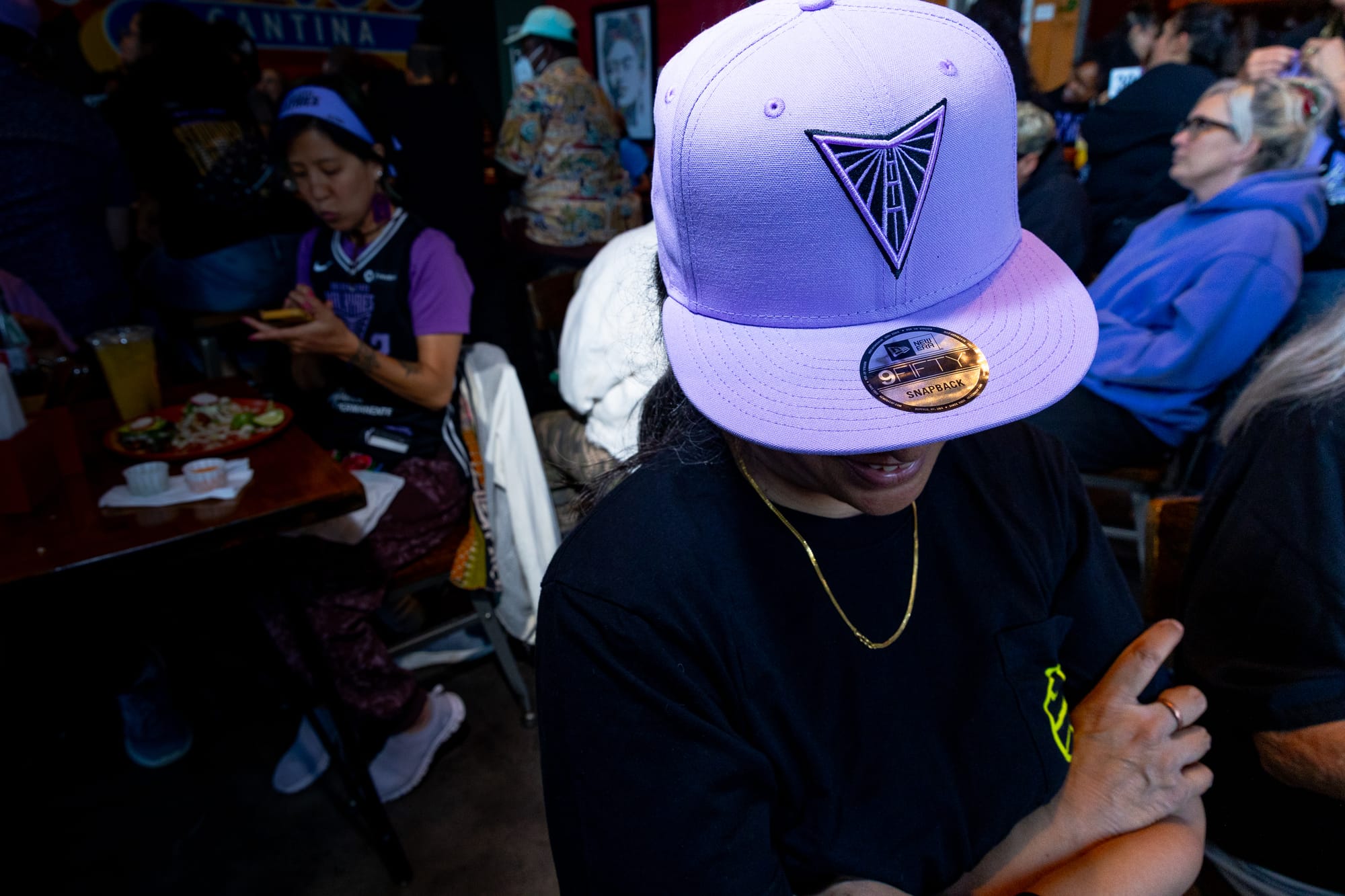 a person stands with their arms crossed in a black shirt with their light purple Valkyries hat obscuring most of their face