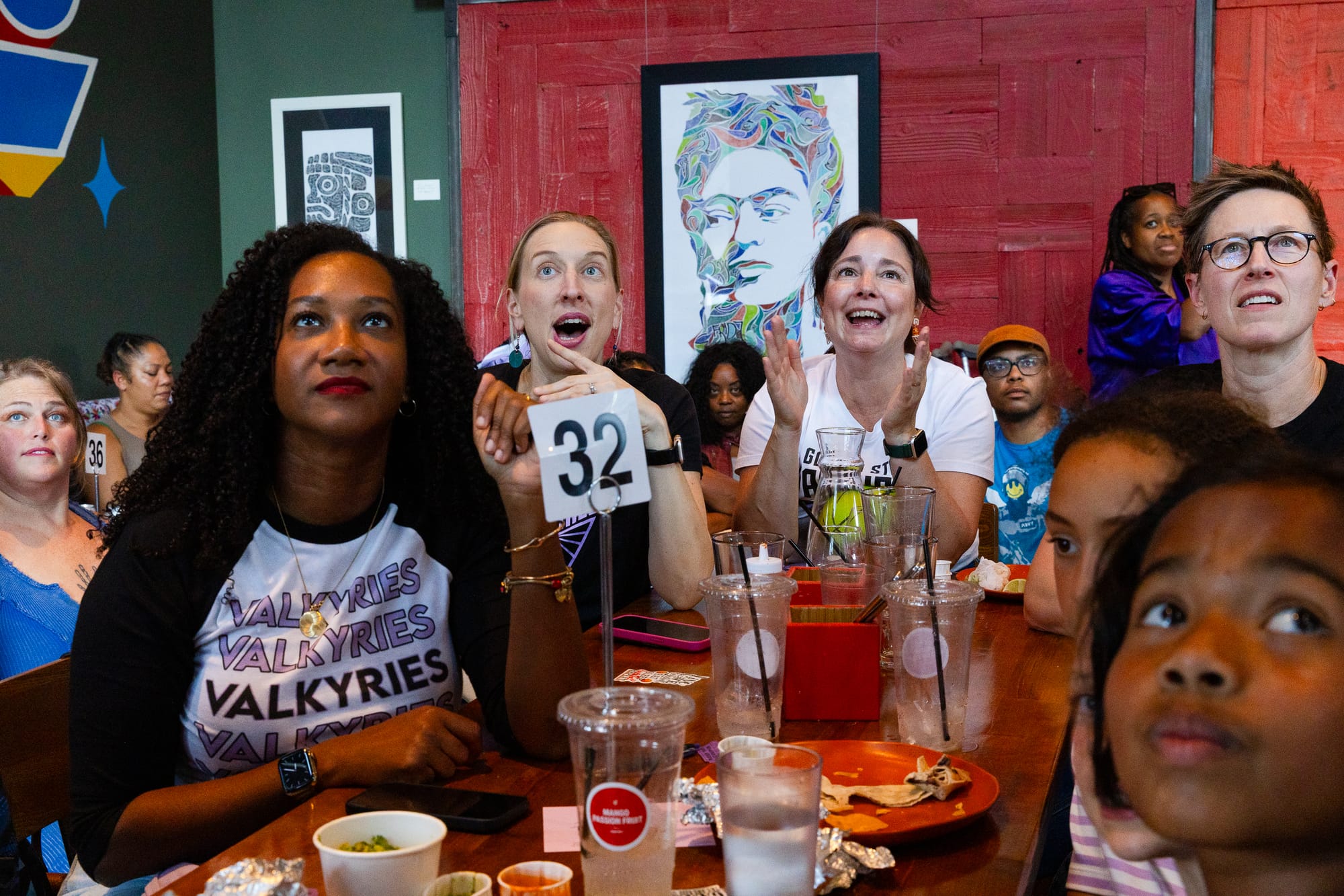 a group of fans of all ages sits around a table in a sports bar looking up at a TV