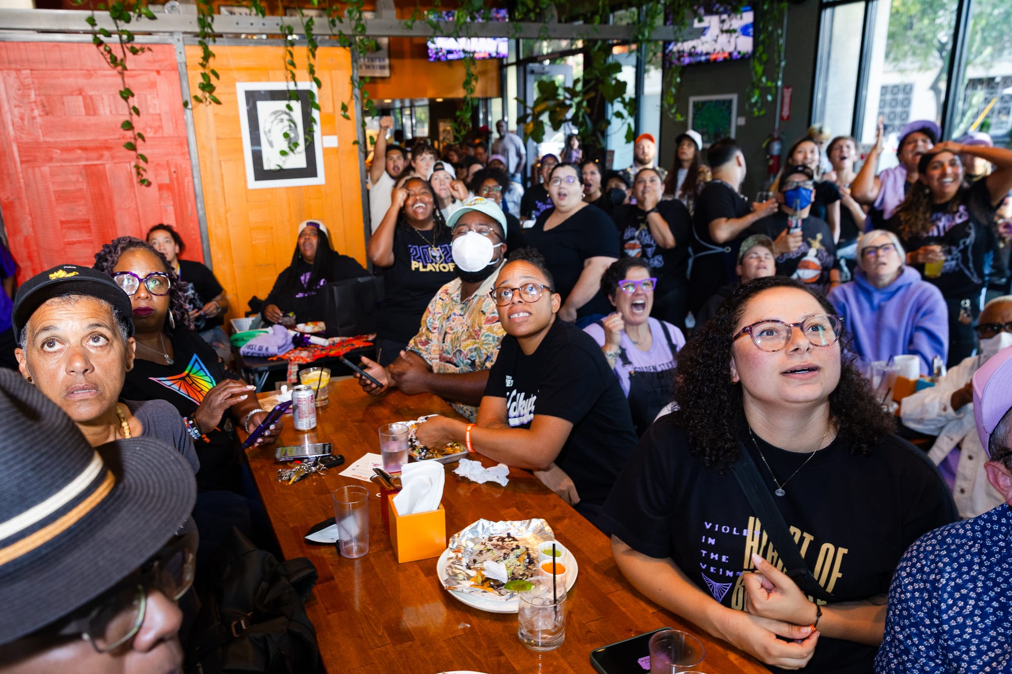 a diverse group of Golden State Valkyries fans wearing purple and black shirts sit around a table at a sports bar, looking up