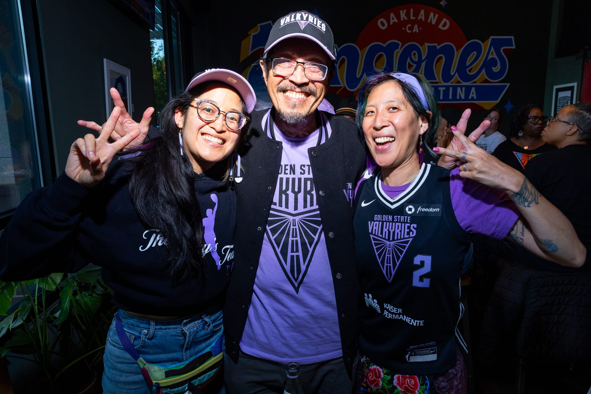 three people wear purple and black Golden State Valkyries gear and pose for the camera with peace signs, two younger people in their late 30s and early 40s with their father, who is in his 70s