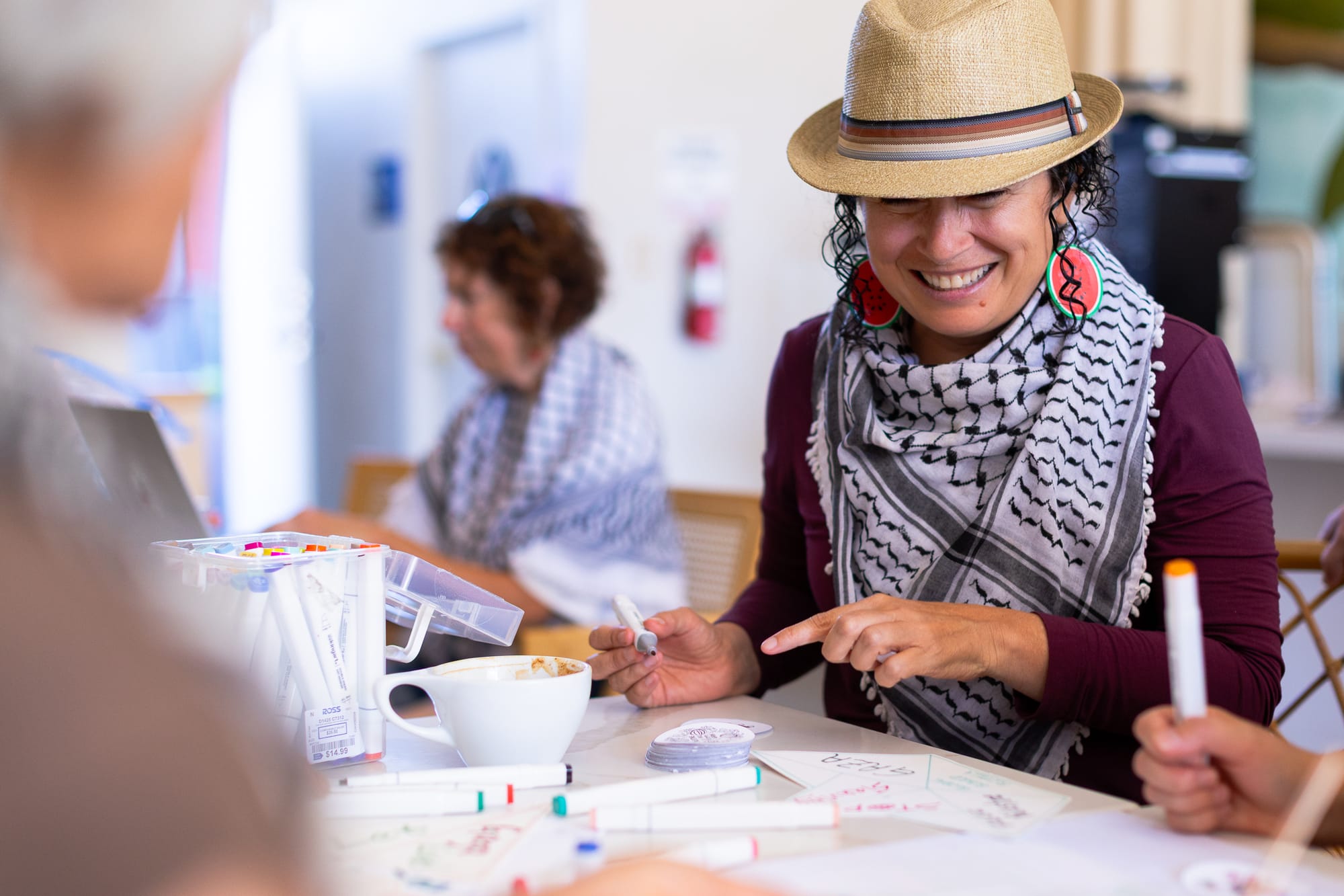 a woman in a maroon top and black and white keffiyah and watermelon earrings smiles while drawing on paper at a cafe