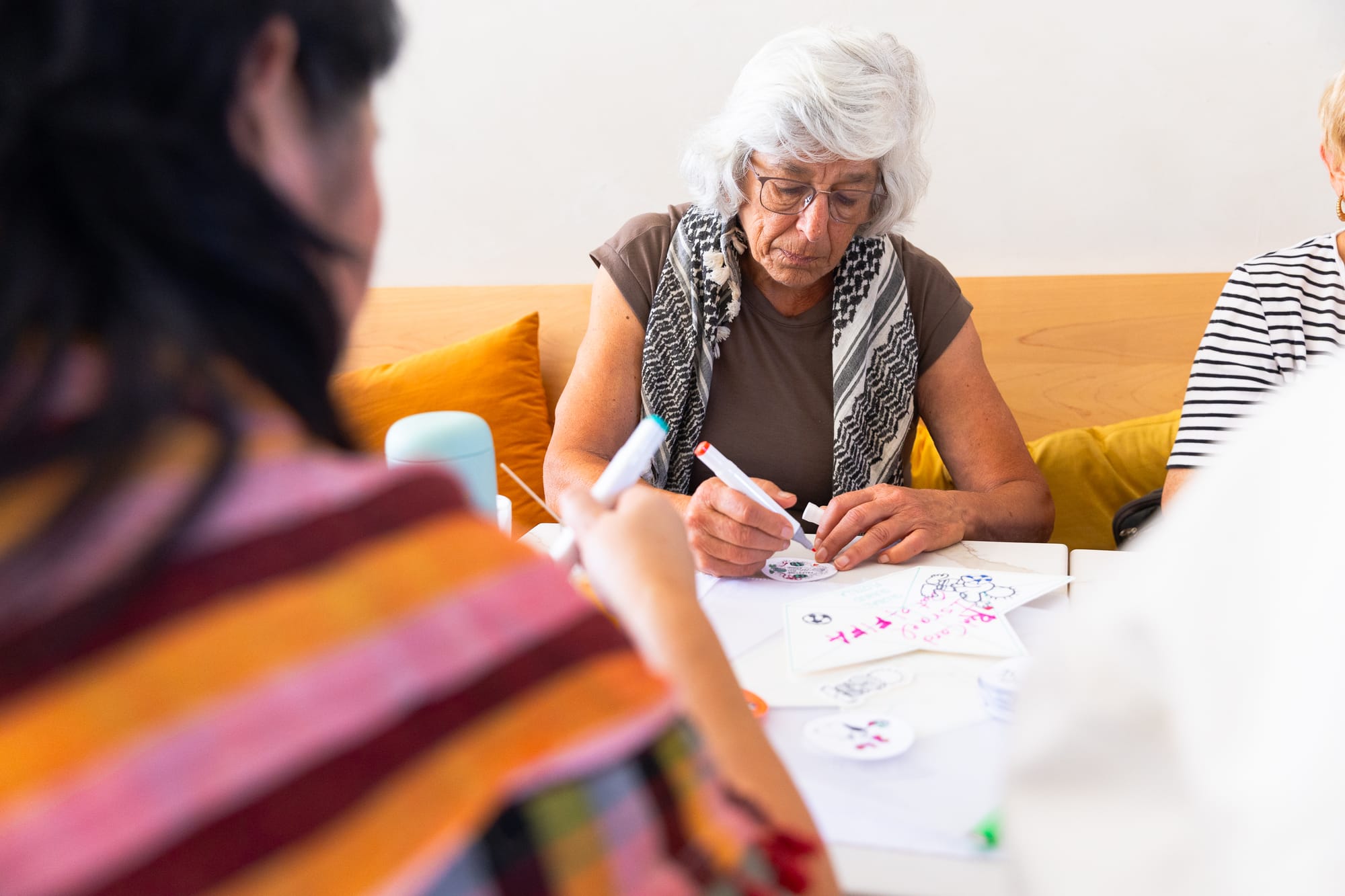 a woman in her 70s sits at a table wearing a keffiyeh, with markers and paper, and two people facing her in the foreground