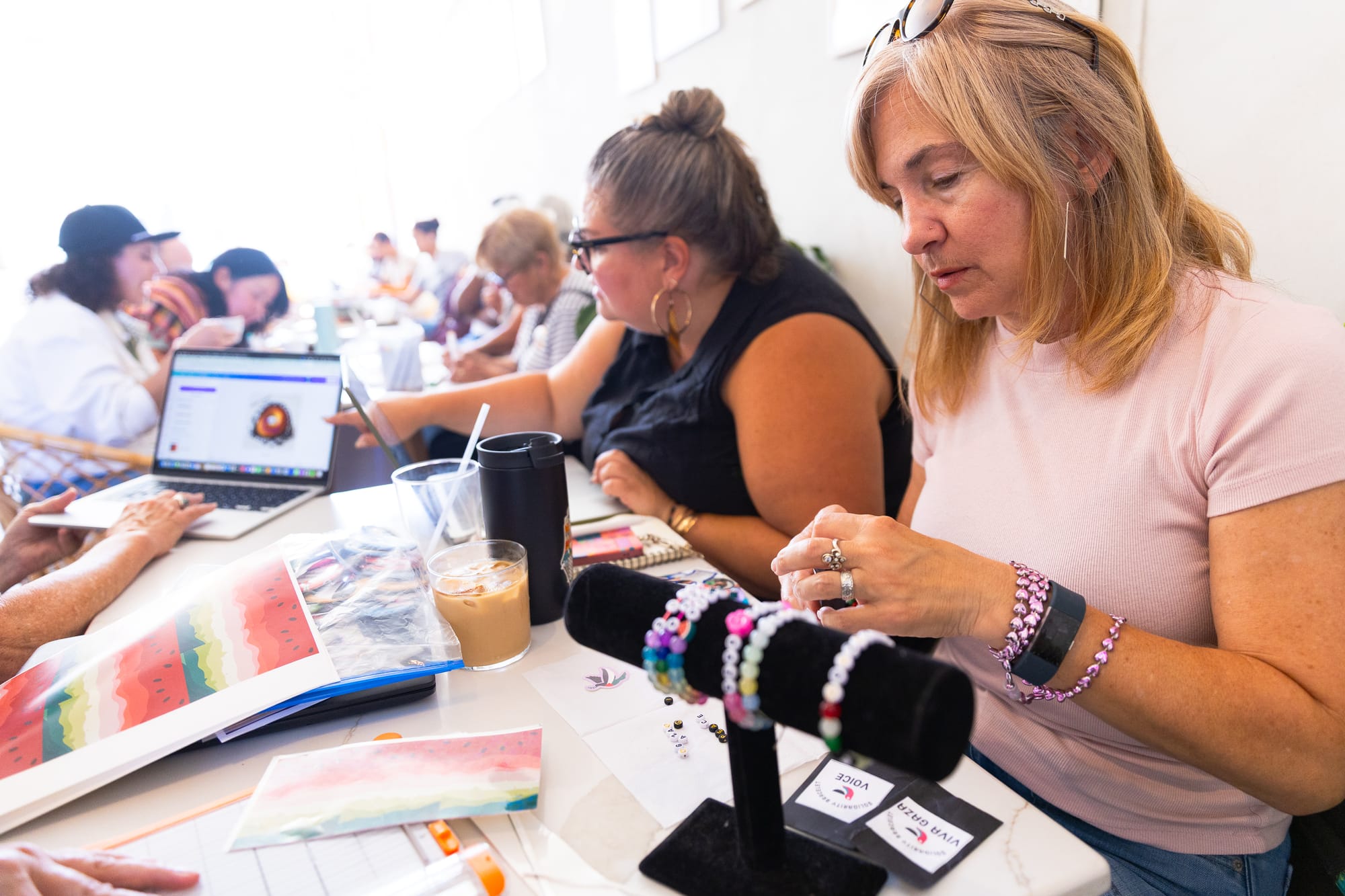 people sit at a crowded cafe doing crafts and talking over books, a woman in the foreground makes friendship bracelets