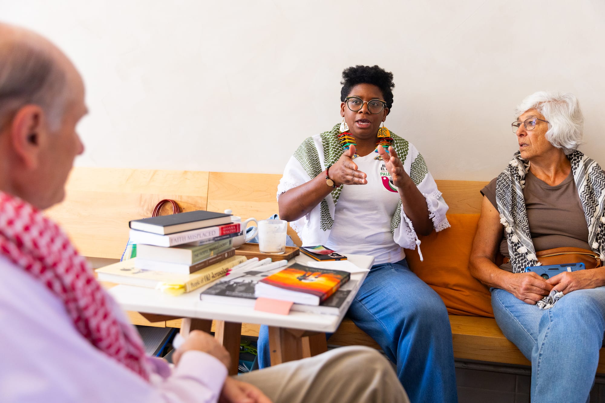 three people sit at a cafe talking in front of a pile of books; two older Palestinian-American people, a man and a woman, and a younger Black woman