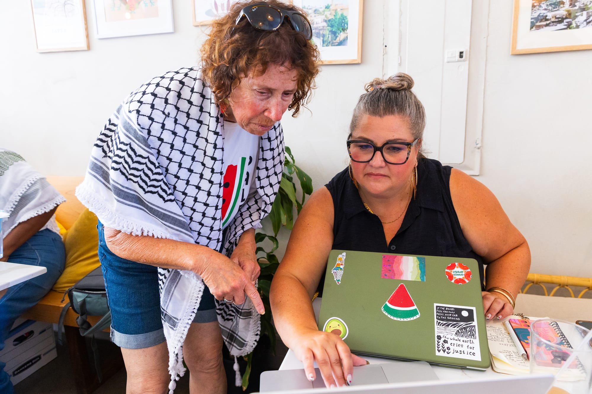 two women look over laptops at a cafe, one is wearing a keffiyeh