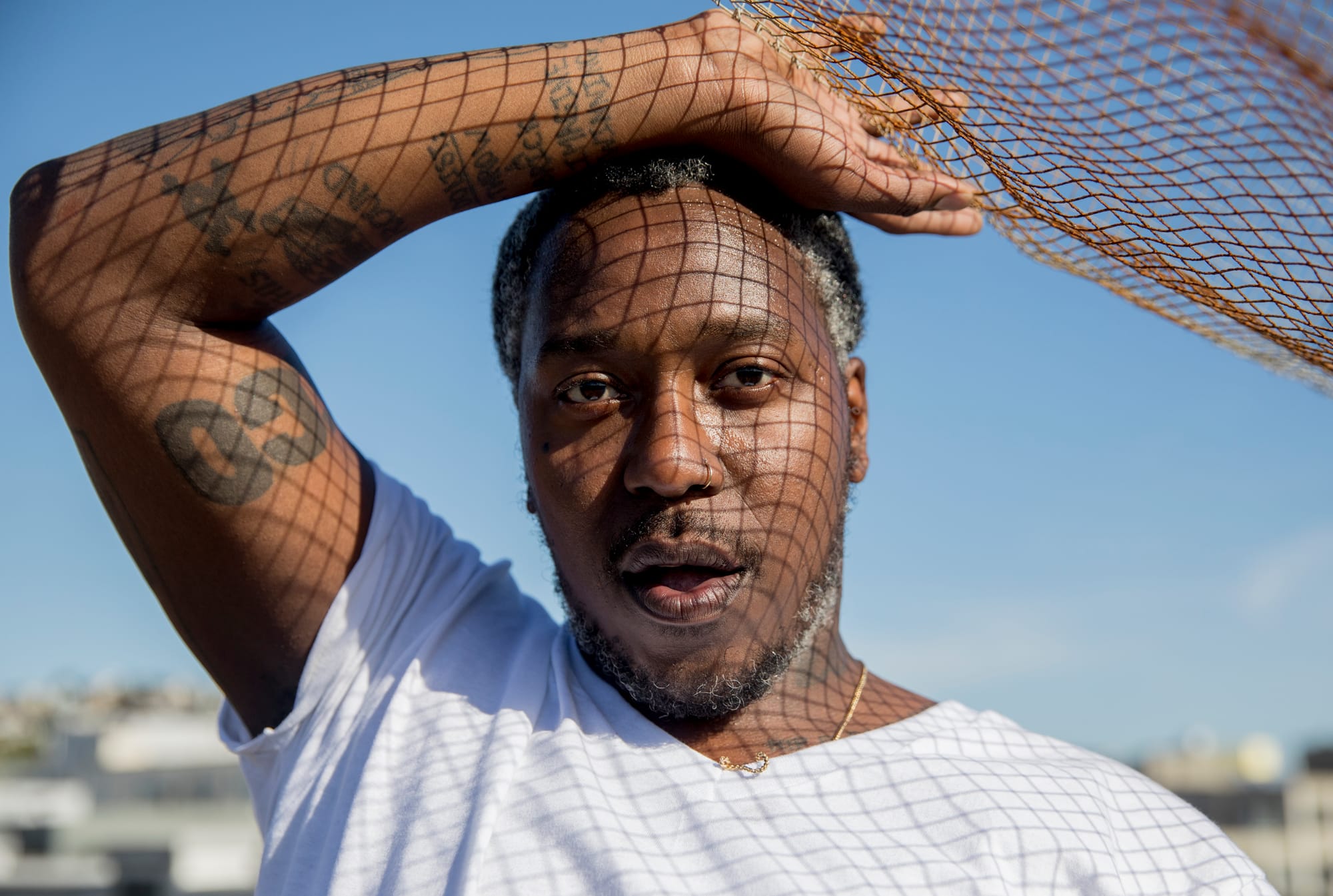 a Black man in a white shirt stands looking into the camera for a portrait against blue sky with his right arm raised over his head