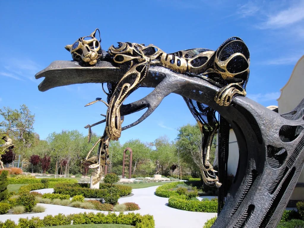 A large metal statue of a leopard sleeping on a branch. The jaguar is gold, its body made of abstracted shapes, with one leg, tipped with claws, dangling down toward the viewer.
