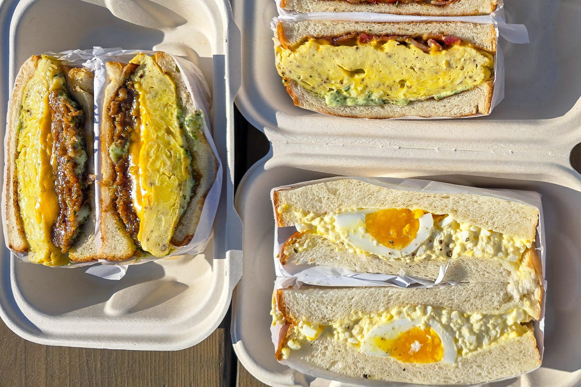 Top-down view of three breakfast sandwiches on white bread. Left: scrambled egg and sausage; right top: scrambled egg and bacon; right bottom: egg salad.