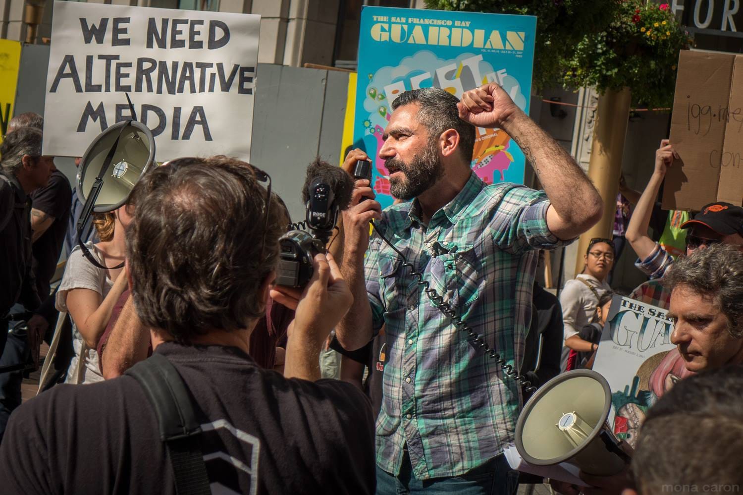 a man in a green and purple plaid shirt speaks into a loudspeaker with his fist in the air at a protest, with people all around holding signs. One reads 'We need alternative media'