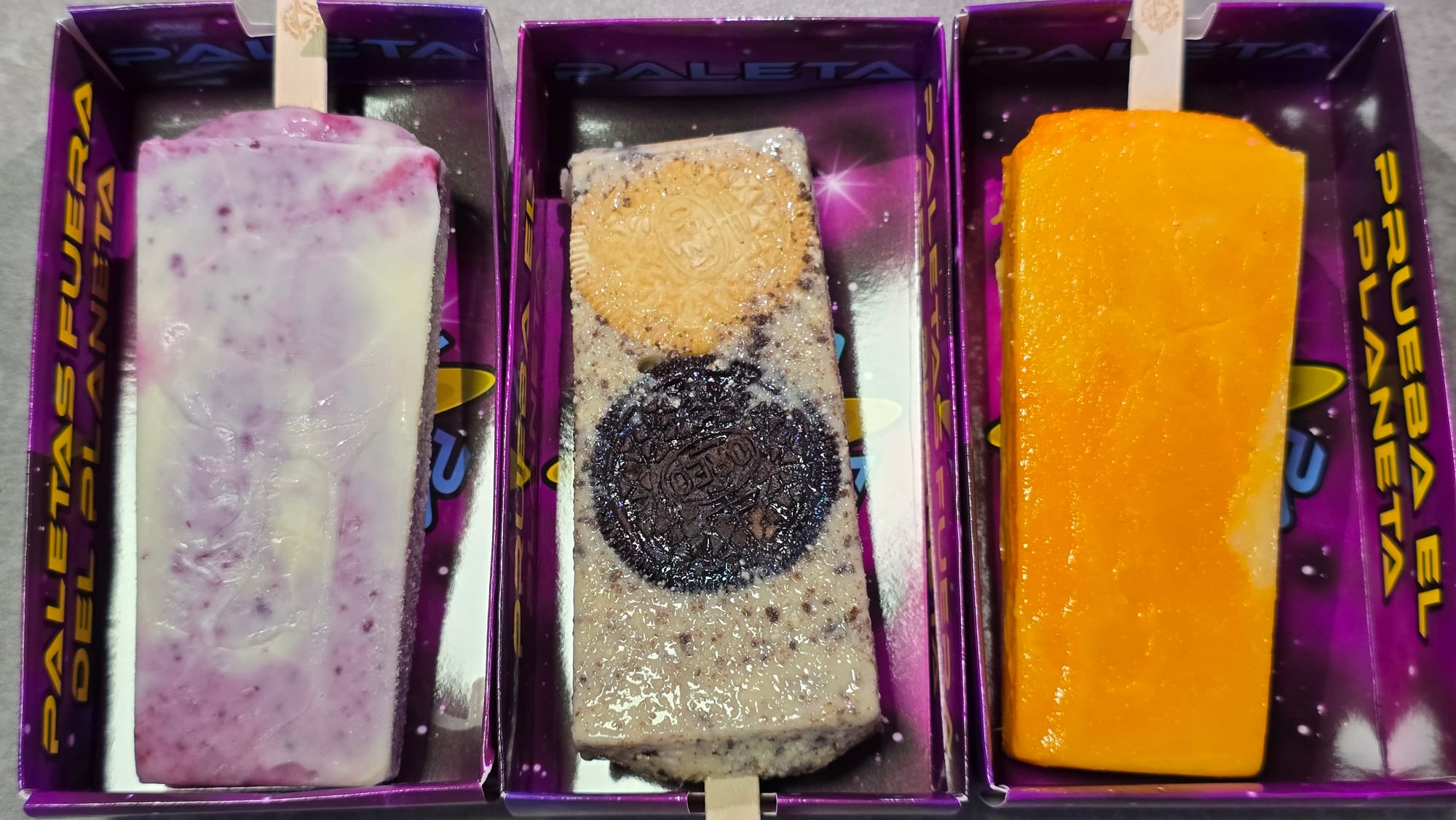 Three paletas in individual purple takeout boxes. Left to right: A purple-and-white paleta, a brown speckled paleta with two cookies inside, and an orange-yellow paleta.