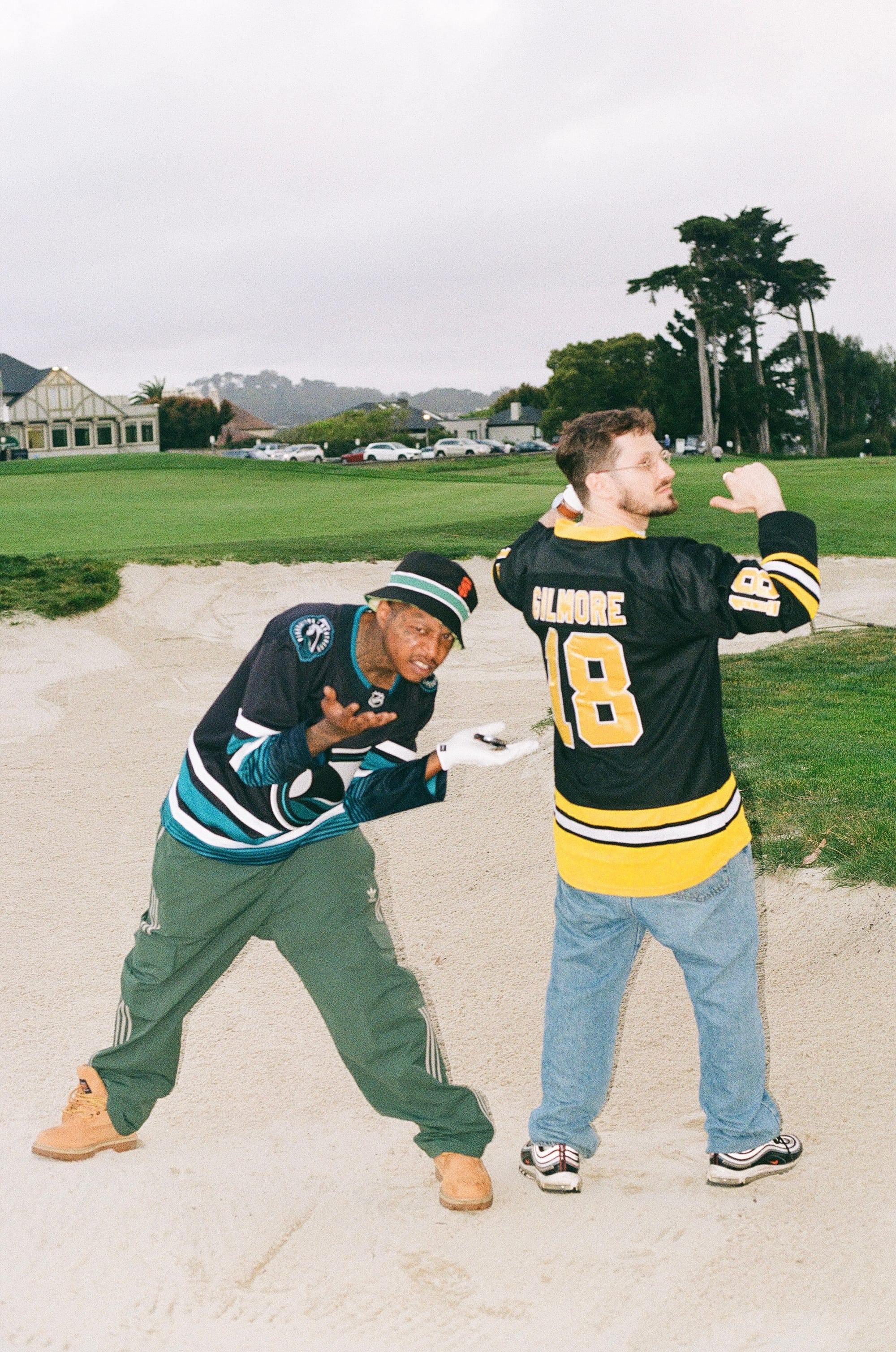 A man in a hockey jersey and green pants stoops in a golf course sand trap. A light-skinned man in a hockey jersey stands with his back to the camera.