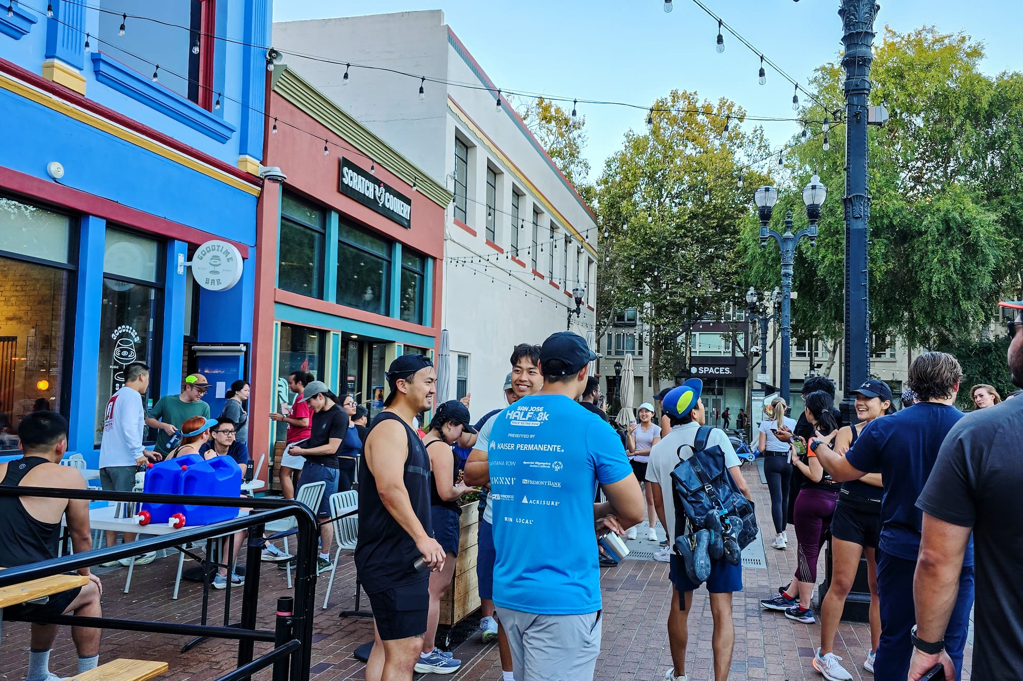 A crowd of people stand in front of a sidewalk patio on an urban street.