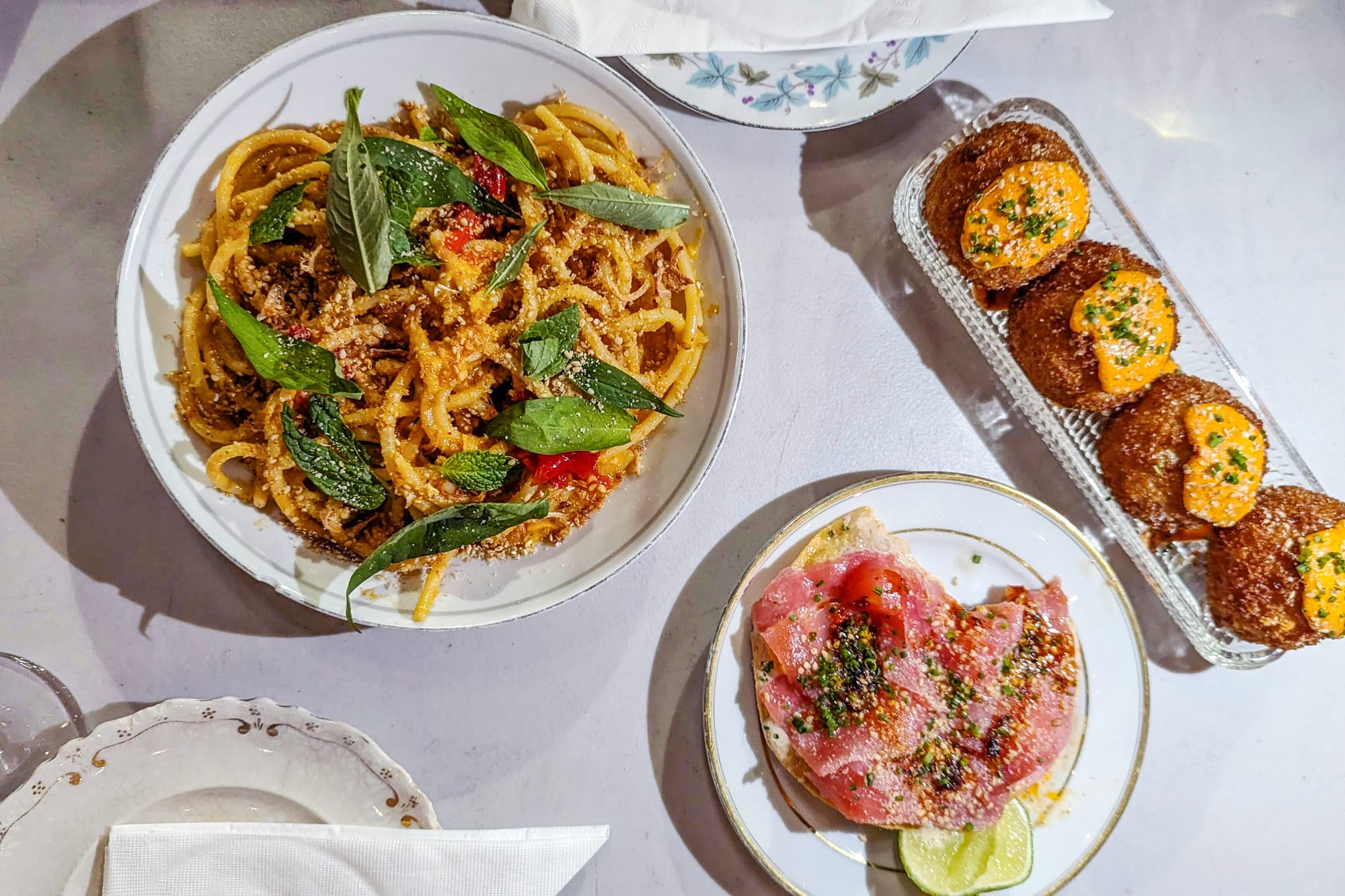 From left to right: A plate of noodles with leafy garnishes, a tostada topped with pink raw tuna, and a plate of four fried balls garnished with uni.