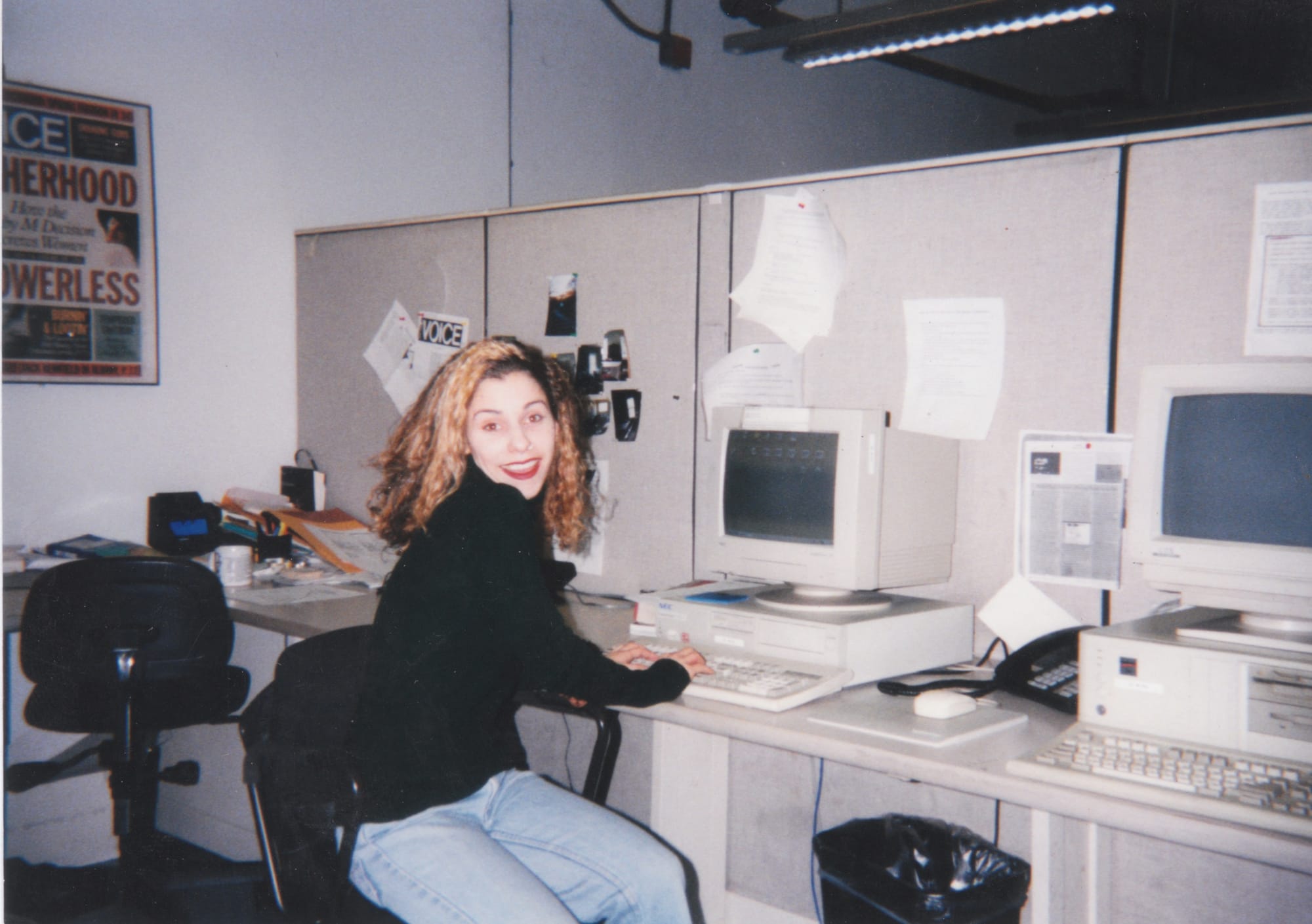 a woman with light blue jeans and a black top and red lipstick sits at a desk with a 90s-looking computer