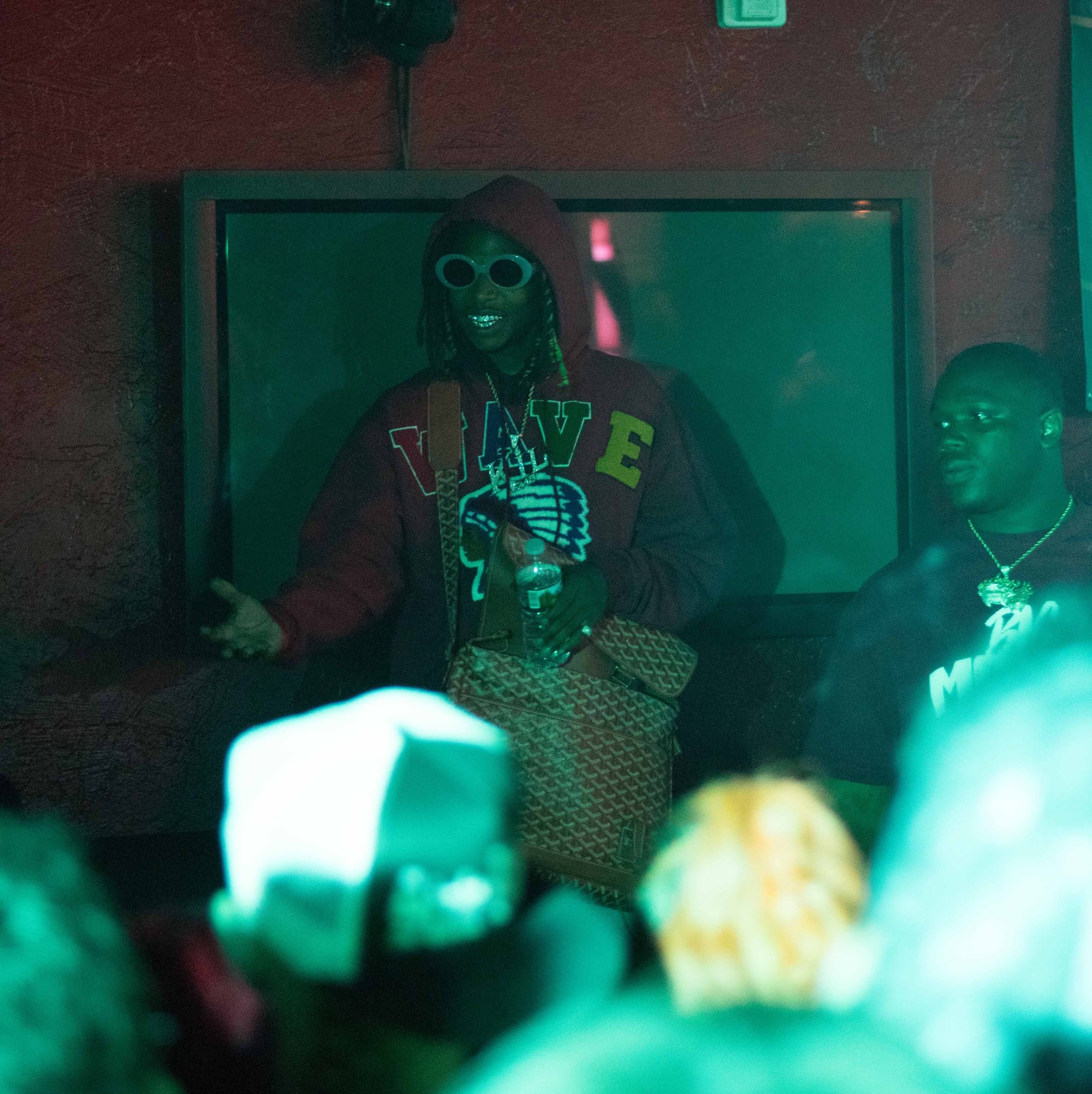 A man in sunglasses and a red hooded sweatshirt that says "Wave" gestures toward a crowd.