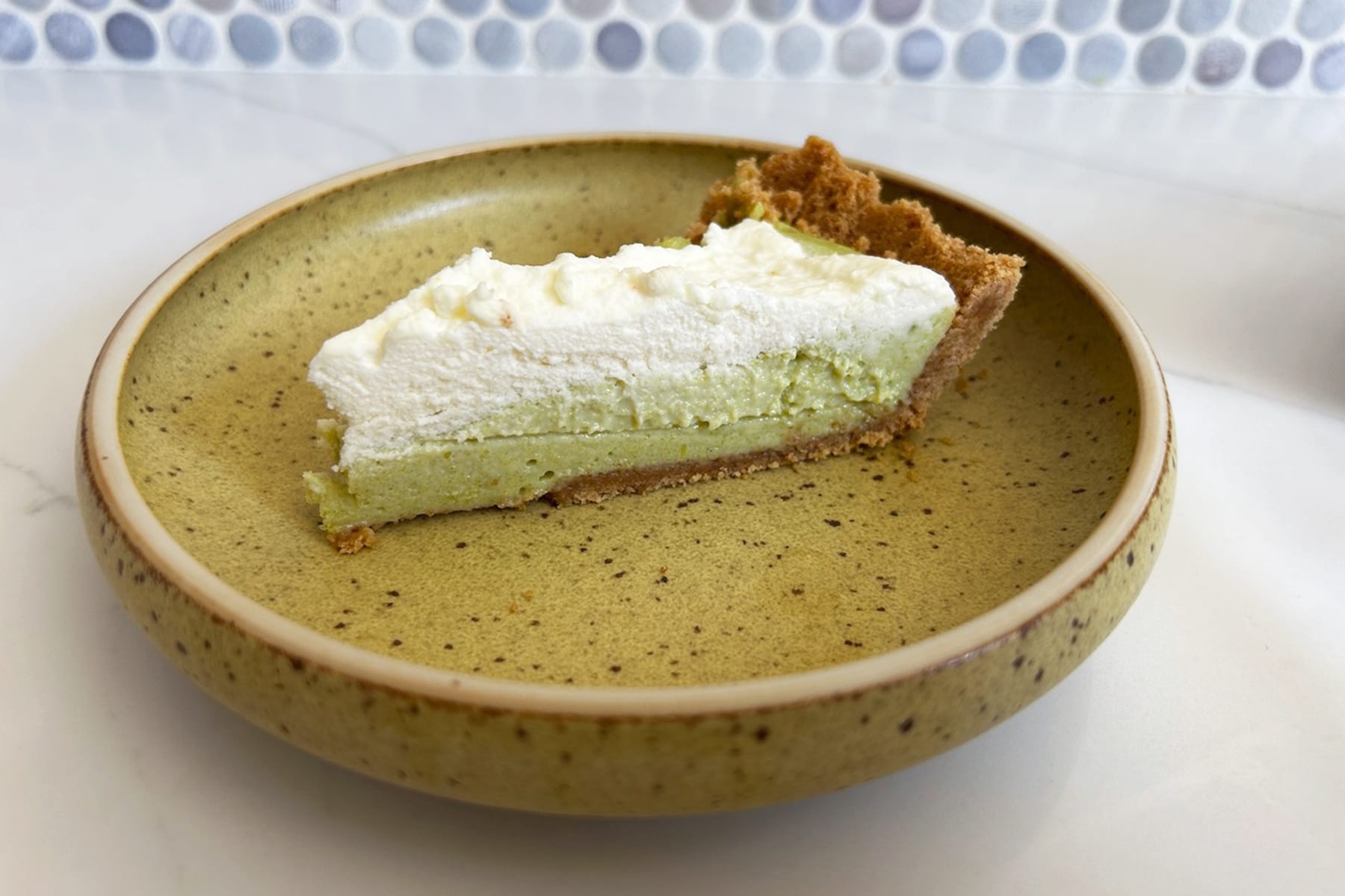 A slice of green pie with white cream on top, sitting on a mustard yellow plate with brown speckles.