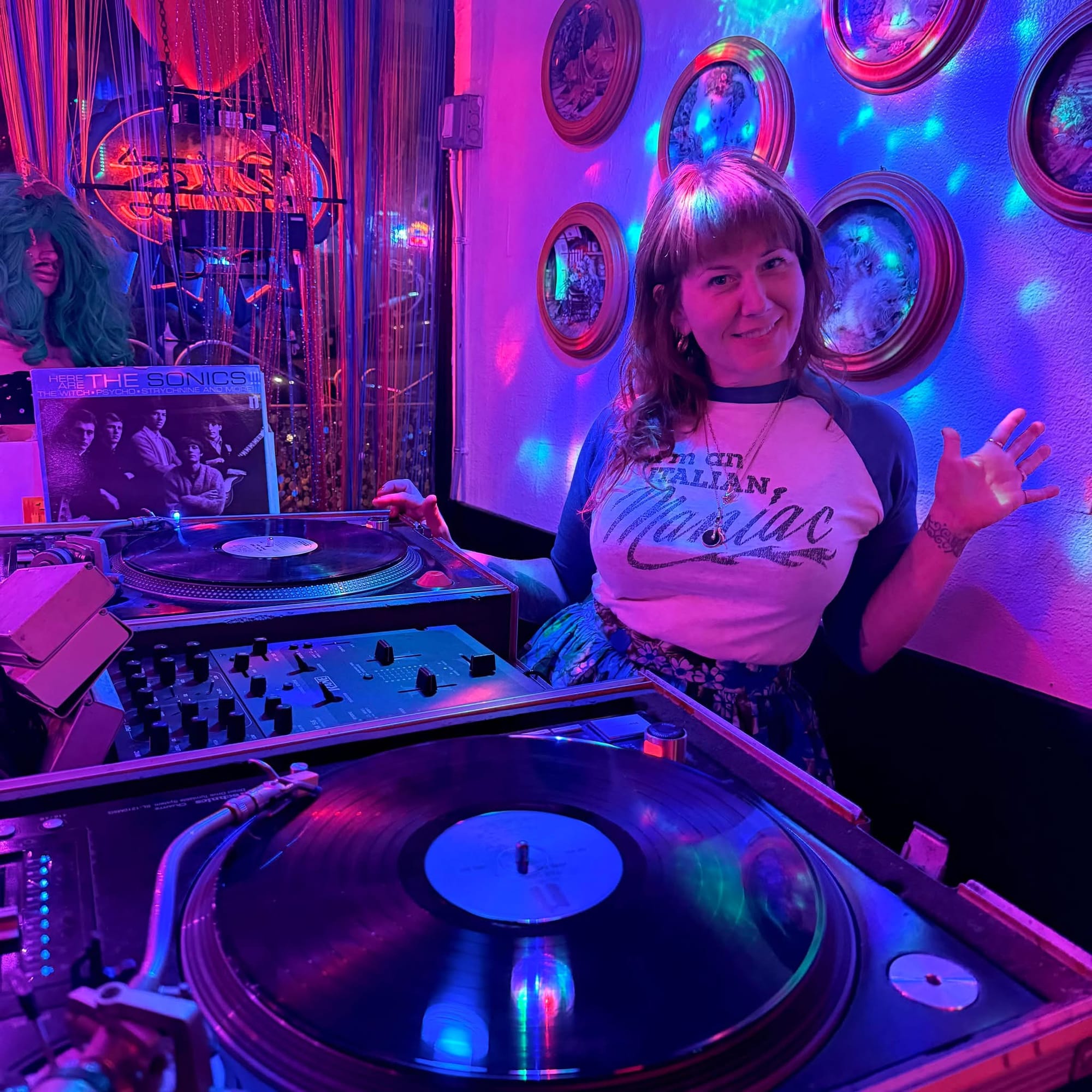 a woman behind a DJ console flashes a peace sign while smiling at the camera