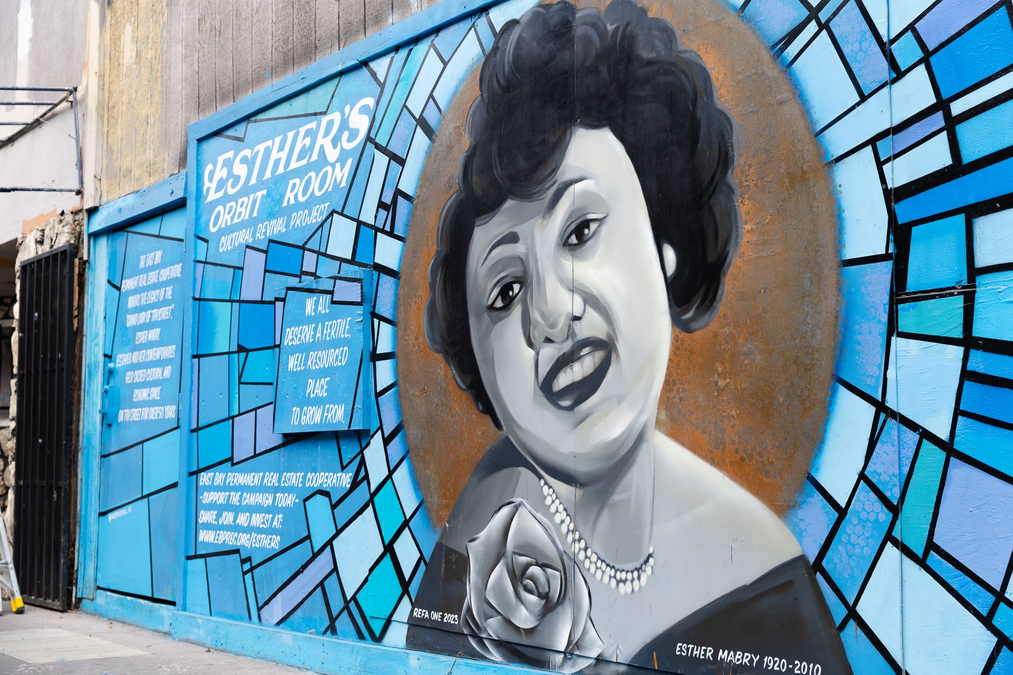 A mural with a black and white portrait of a woman in the middle. She has a rose, and a pearl necklace. Around her is a blue design, that looks like stained glass. The text says "Esther's Orbit Room cultural revival project. We all deserve a fertile, well resourced place to grow from."