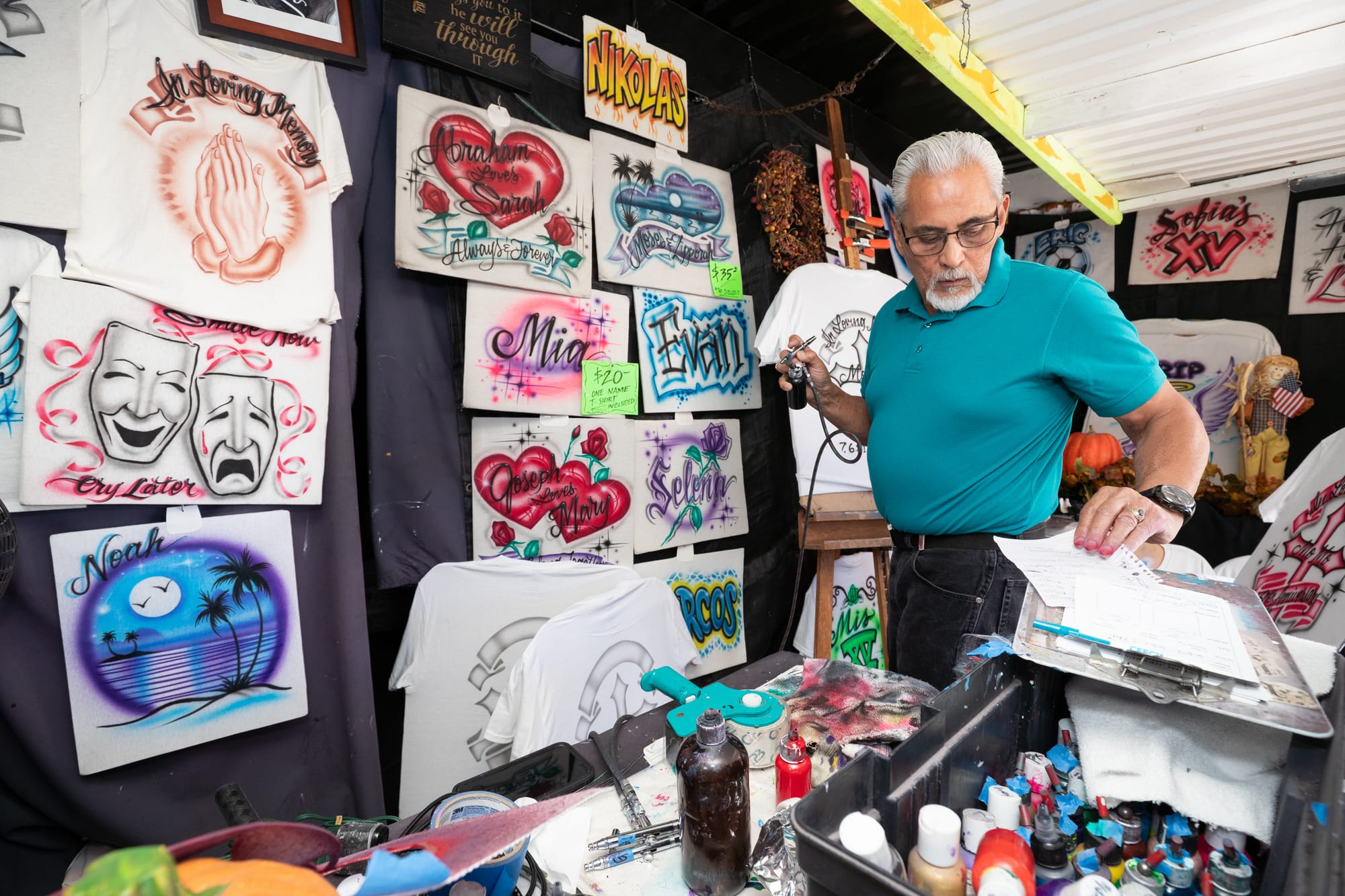 Everett Regua consulting an order form while airbrushing in a tent filled with airbrushed work.