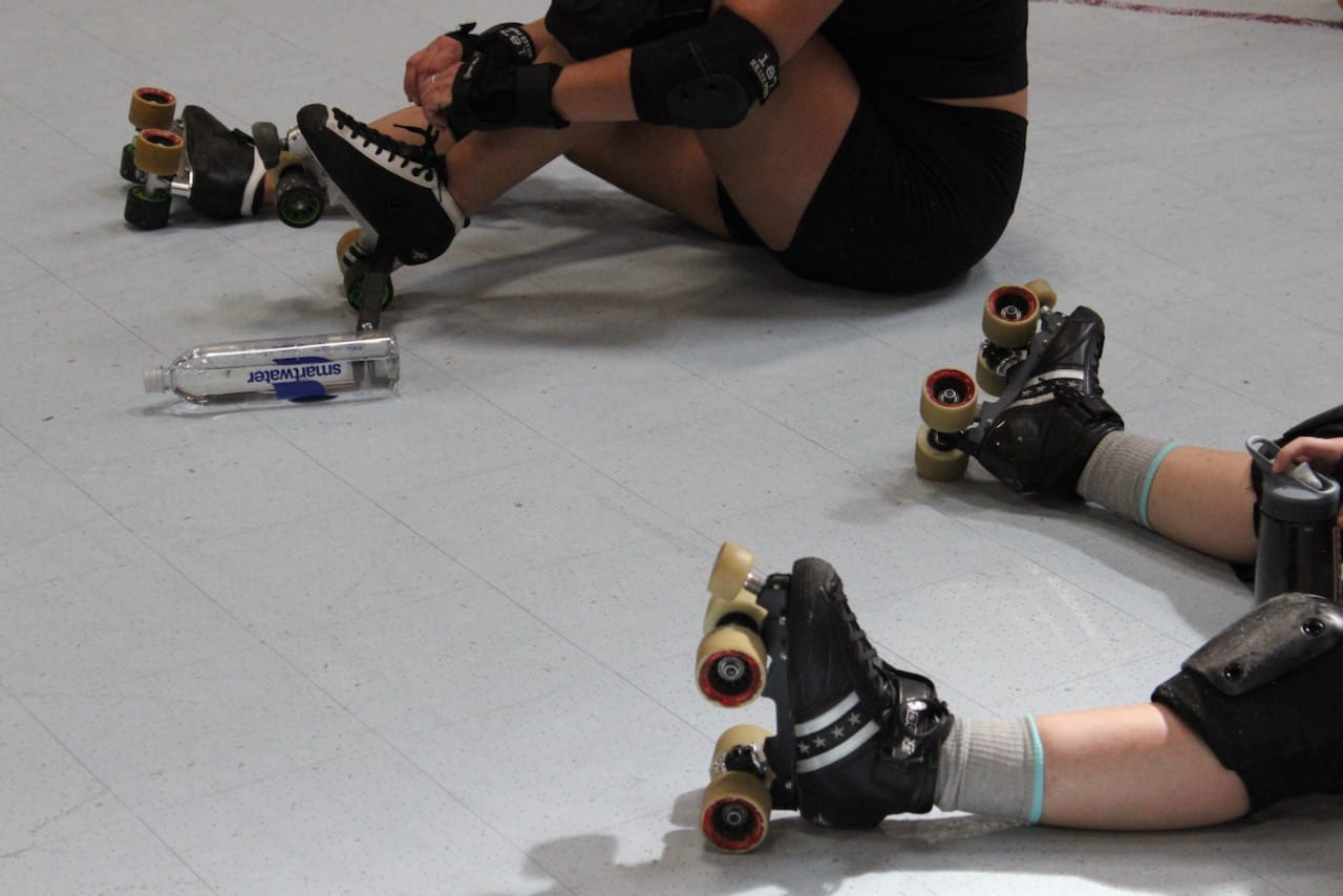 the legs of two skaters, sitting on the ground between drills at practice
