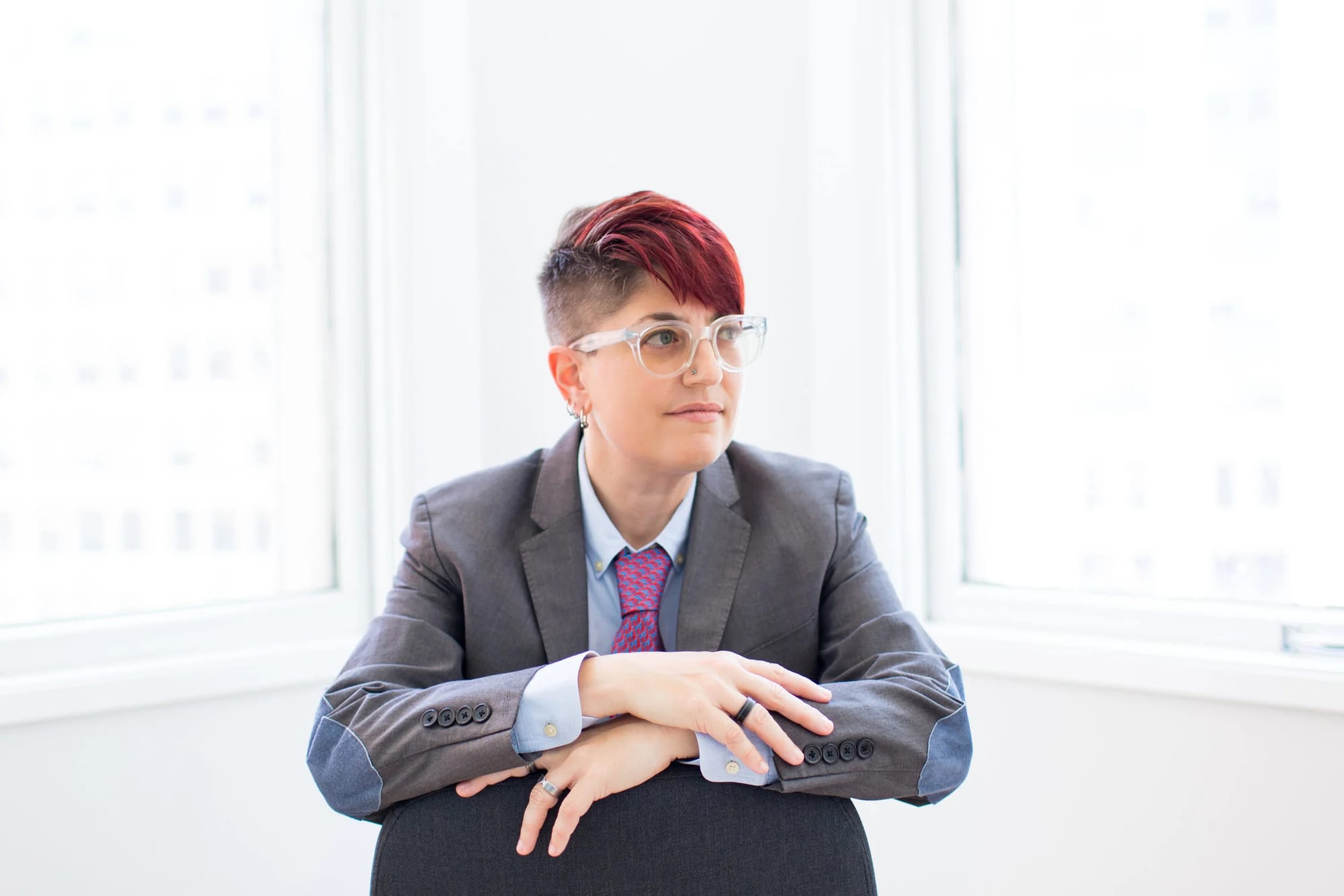 a person with glasses and short bright red hair in a suit sits for a portrait, looking off to the side