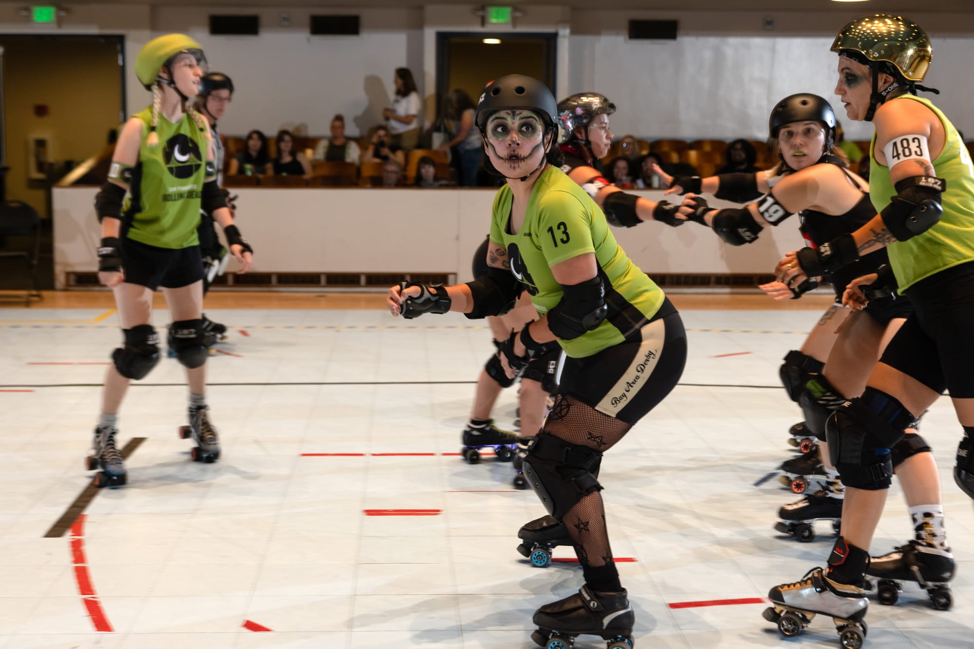 A skater with a green jersey and face makeup to look like a calavera is looking just off camera. Around them are other skaters, mid push and pull.