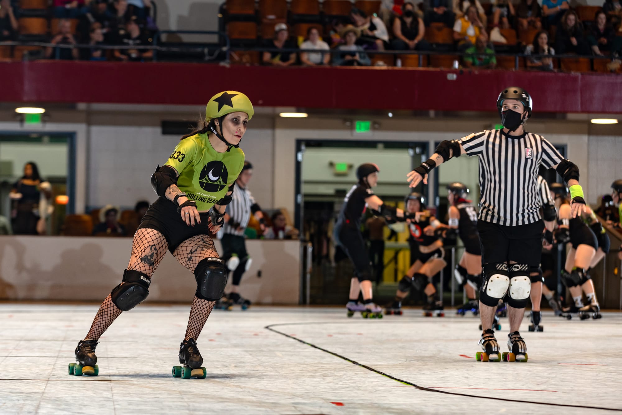 A skater in a green jersey and black fishnets skates around the track, while a referee with a black and white striped jersey watches.