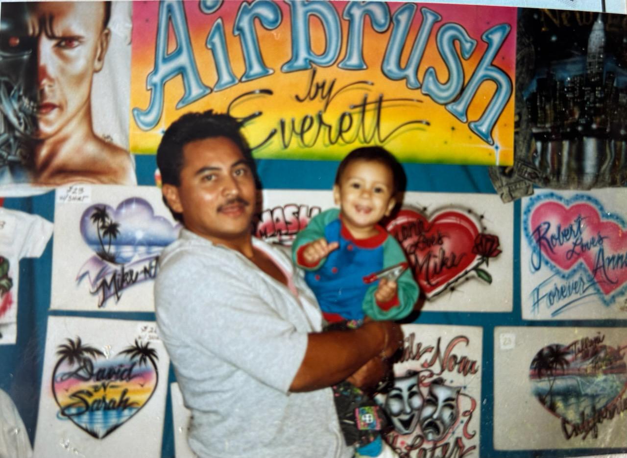 Everett Regua holds his toddler-age son in front of his airbrushing oeuvre.