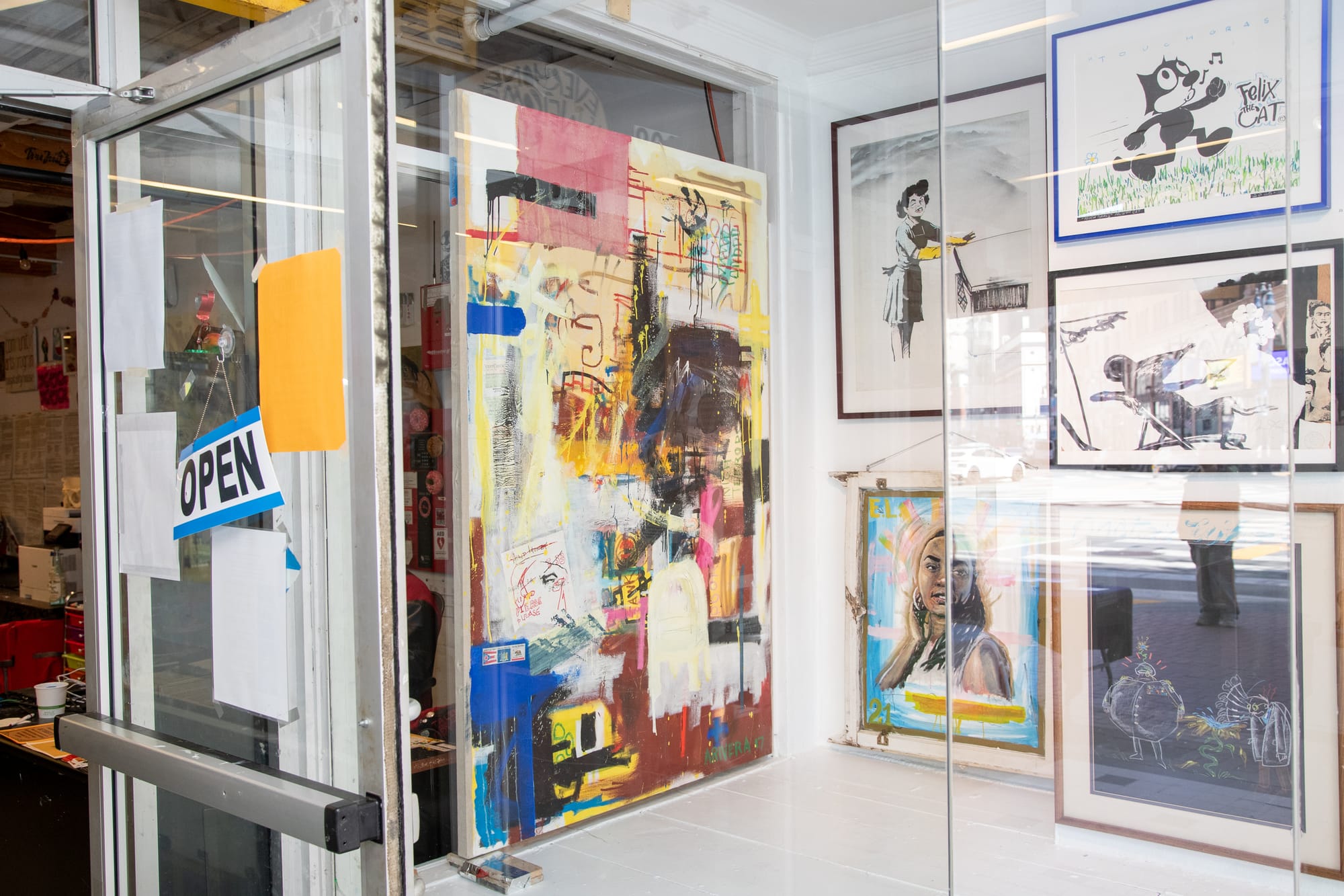 A large painting, titled "Breaking Point", is hung against the wall of a gallery, next to a door with an "open" sign hanging on it. The painting is abstract, with yellow, cream, blue, and pink paint.