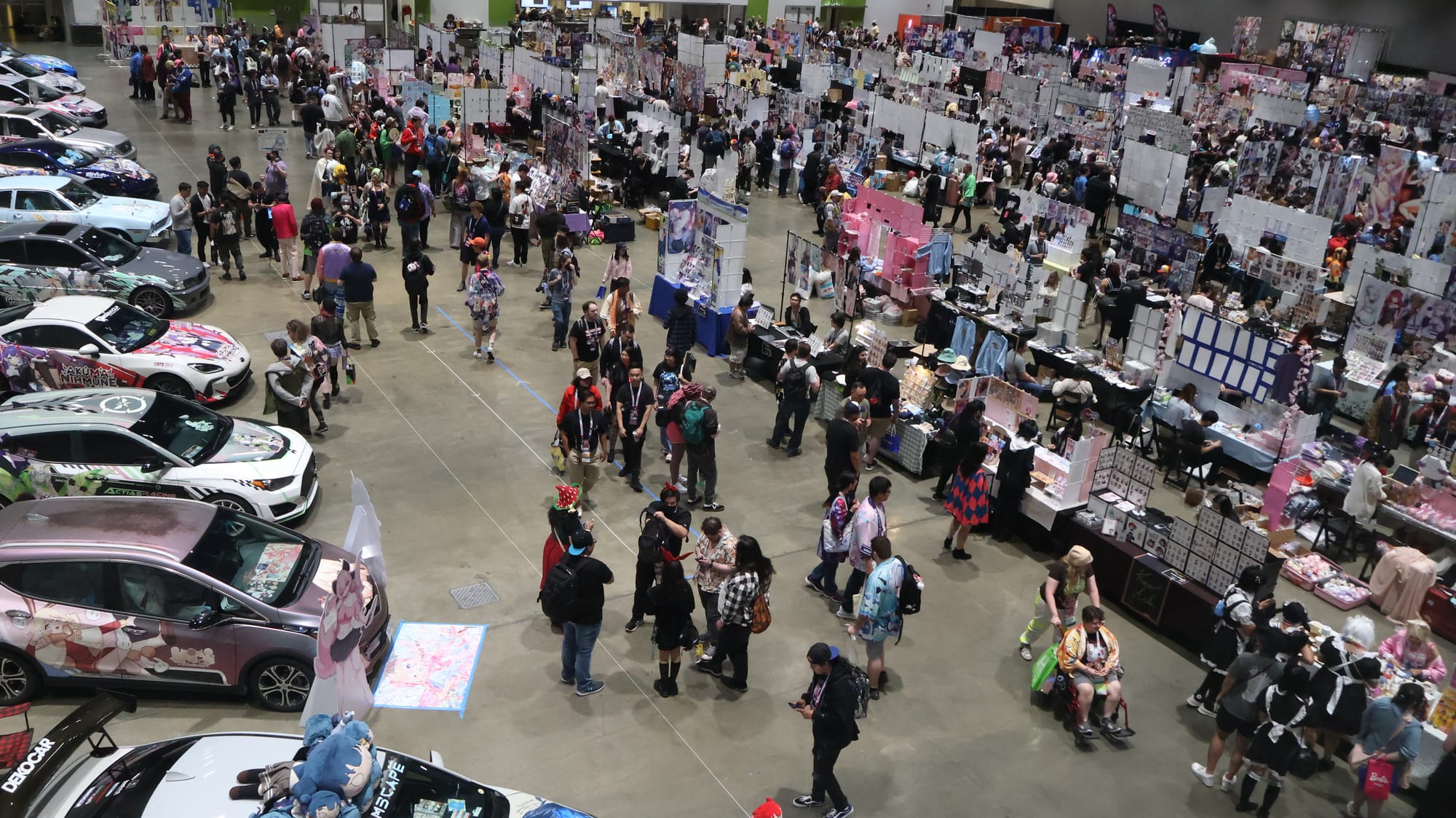 A crowd of people linger in a convention hall with cars wrapped with anime designs and numerous stalls with art displays.