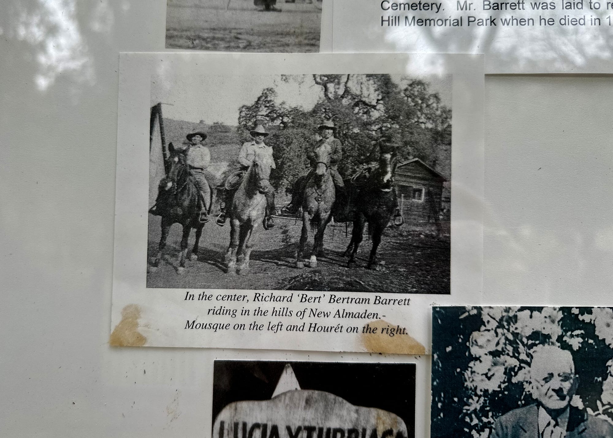 Black-and-white archival photos taped to a display wall. One depicts four horses and three men. A caption reads, "In the center, Richard 'Bert' Bertram Barrett riding in the hills of New Almaden - Mousque on the left and Houret on the right.