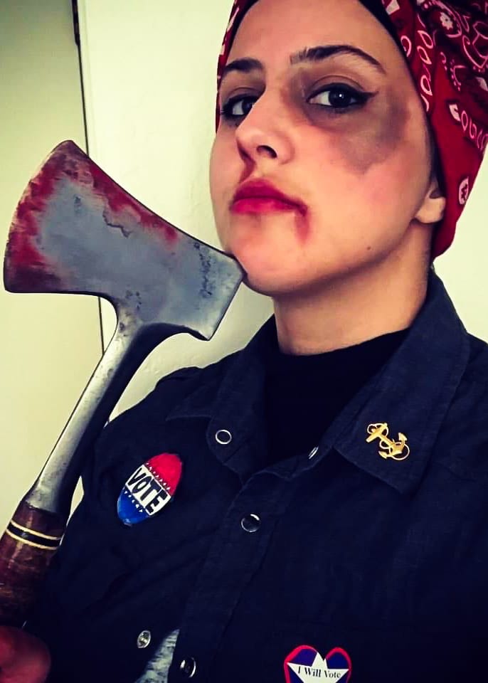 A woman wearing gory makeup, a red handkerchief in her hair, holds an axe.