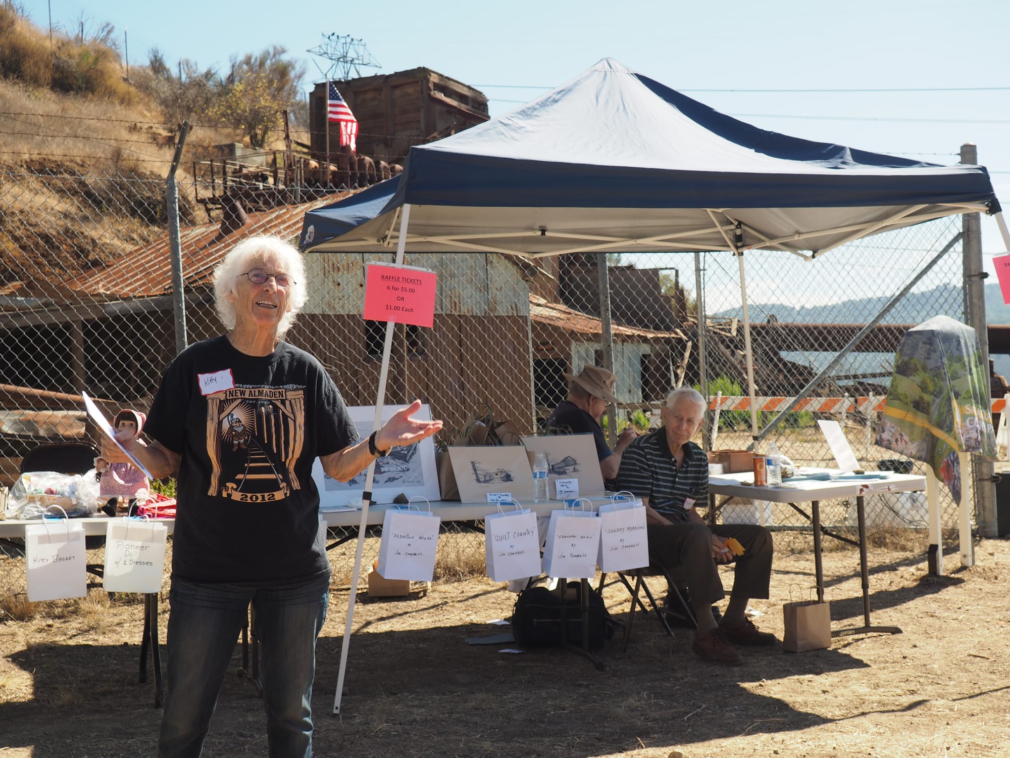 An older woman with white hair speaks in front of an outdoor stall.