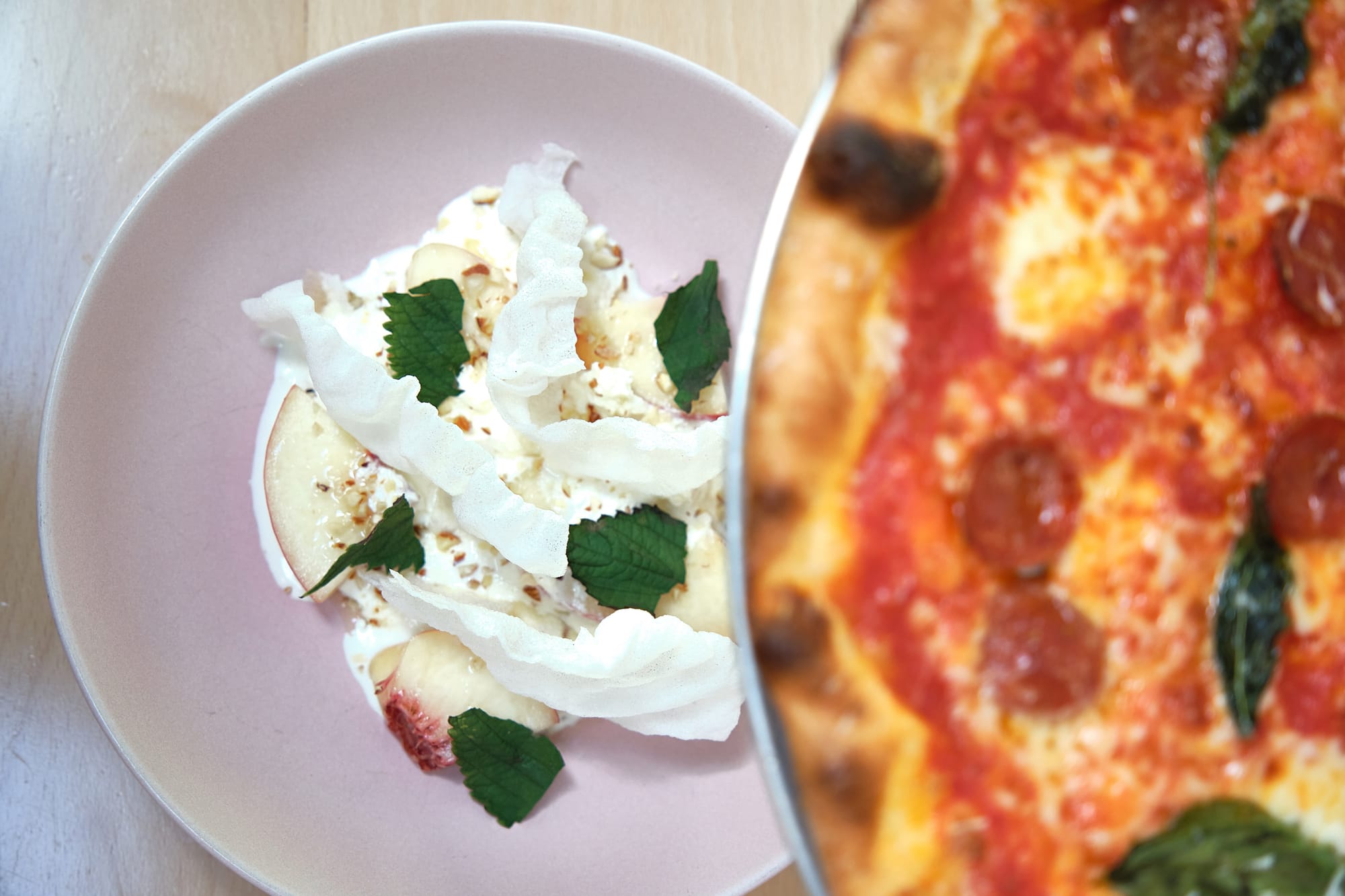 A peach and burrata salad on a white plate under a pizza on a tray.