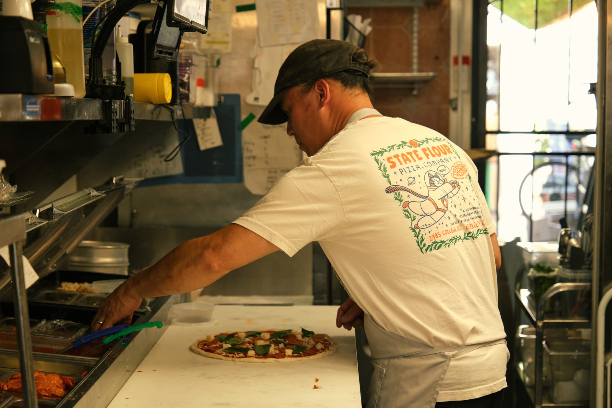 A man in a white t-shirt with a space cat illustration on the back puts toppings on a pizza in a large kitchen.