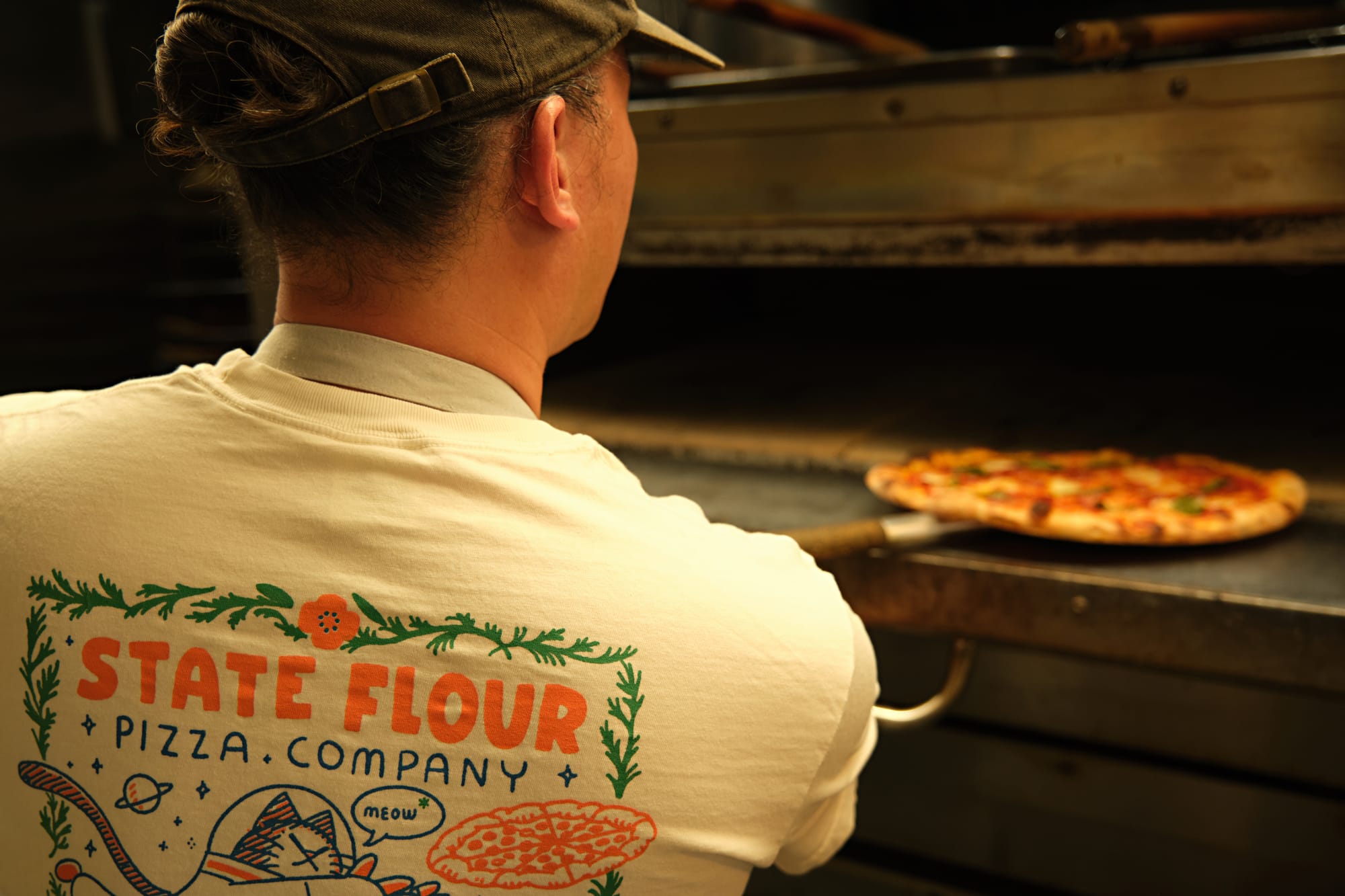 A man in a white t-shirt with a space cat illustration on the back puts a pizza into a pizza oven.