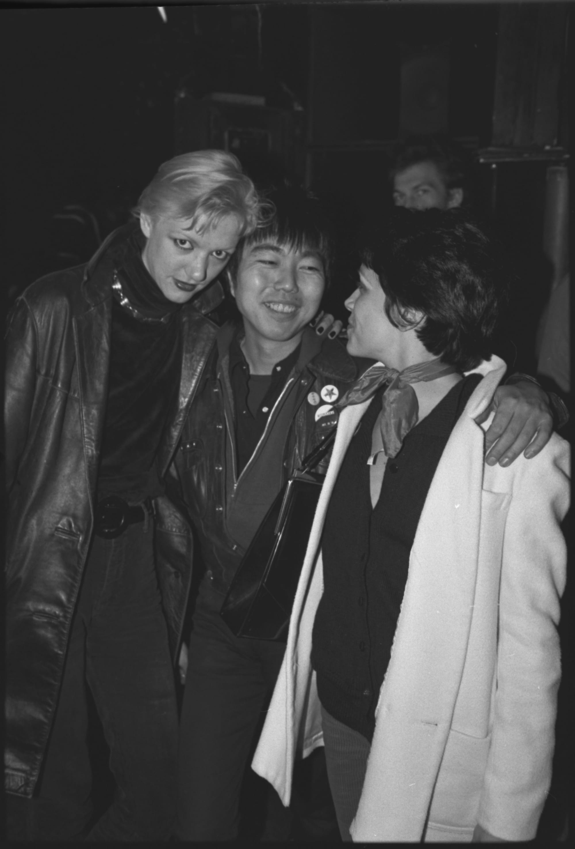 three people, a blonde woman, an Asian man, and a dark-haired woman smile in a black and white photo