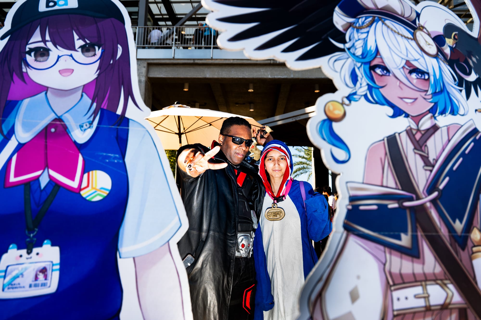 Two people pose behind two cardboard standees of a purple-haired anime girl and a white-haired anime boy.