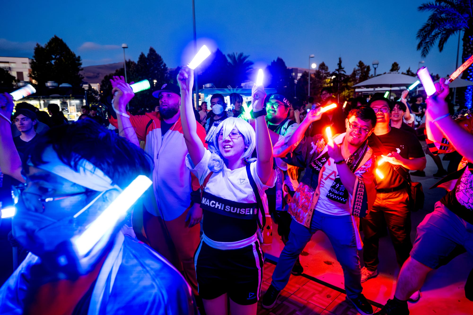 A group of people holding glowsticks dance in a parking lot.