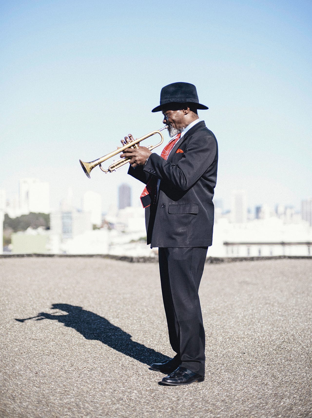 a short Black man in a black suit plays the trumpet, in profile, standing on a gravel roof with a city skyline behind him