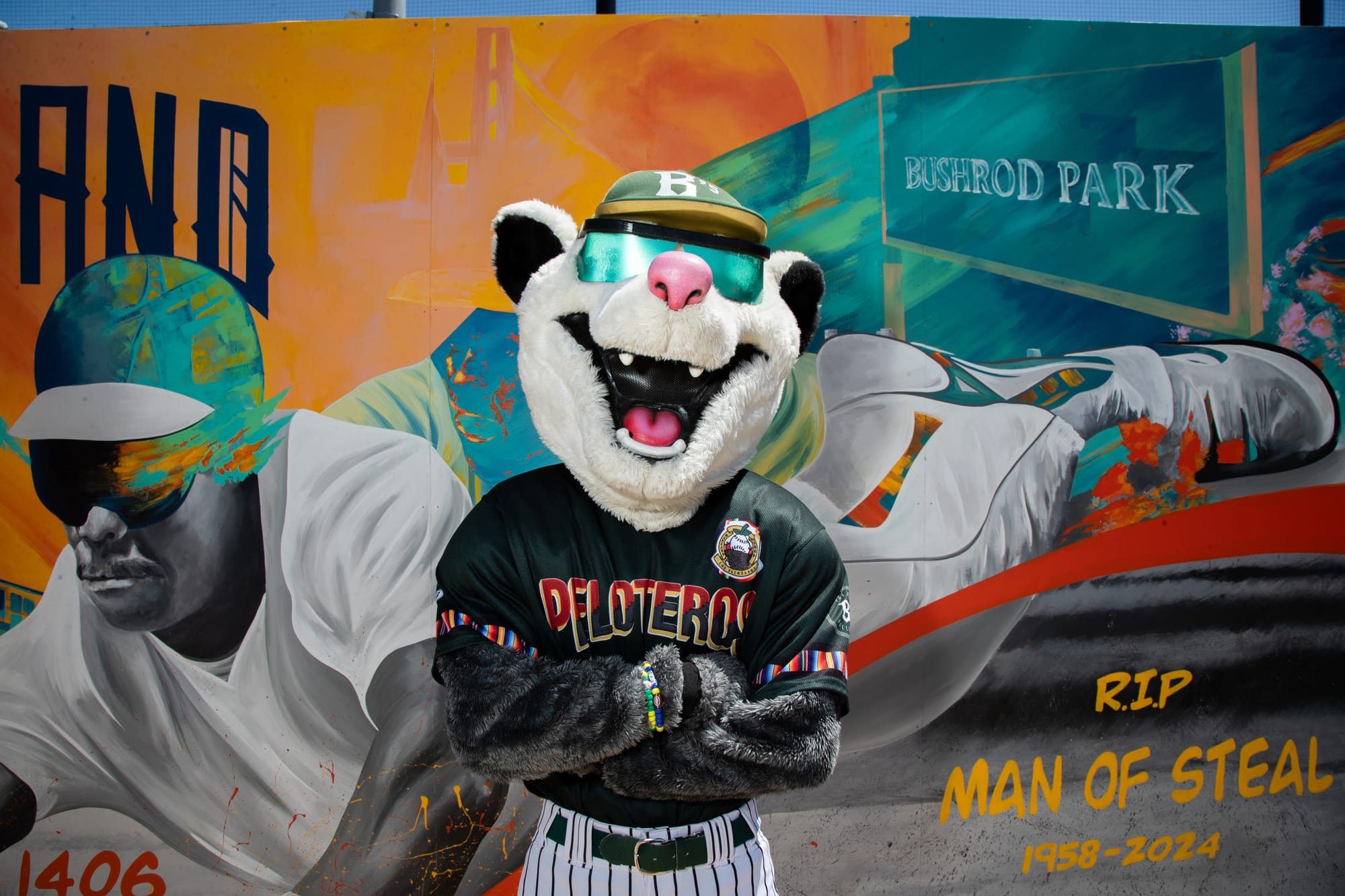 A white possum mascot stands in front of the camera, with his arms crossed looking cocky. He has green sunglasses on and a jersey that says "peloteros" on it.